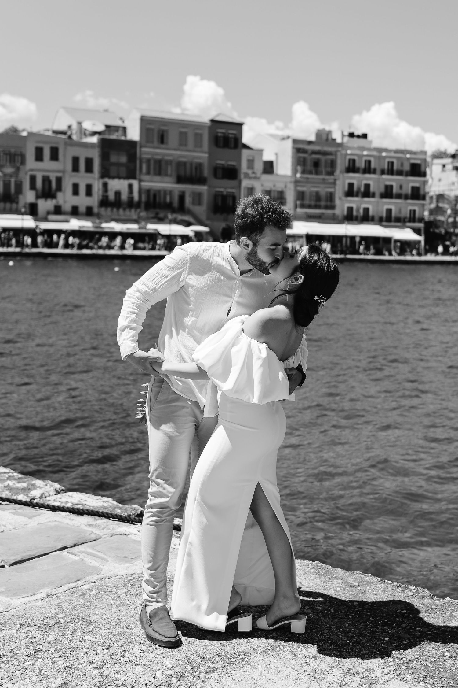 Black and white photo of newlyweds sharing a passionate kiss on a Mediterranean harbor dock with historic buildings behind.