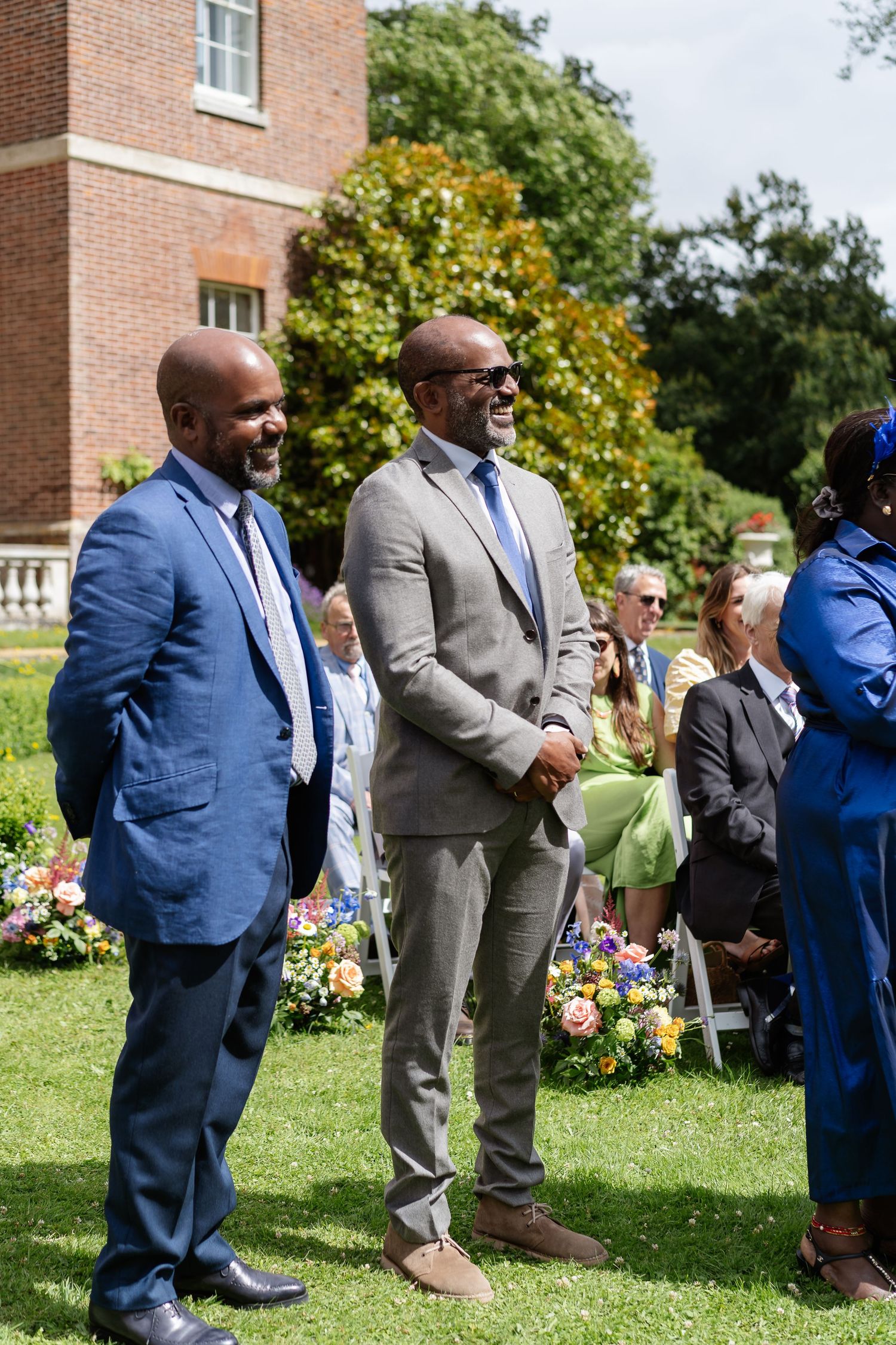 Two smartly dressed male wedding guests smiling and laughing during outdoor ceremony at brick manor.