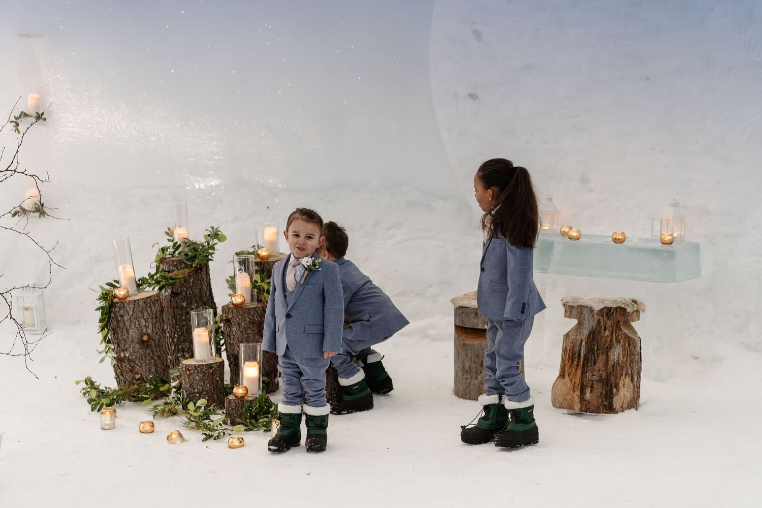 Two children pose in a magical ice cave setting with candles and wooden stumps decorated with greenery.