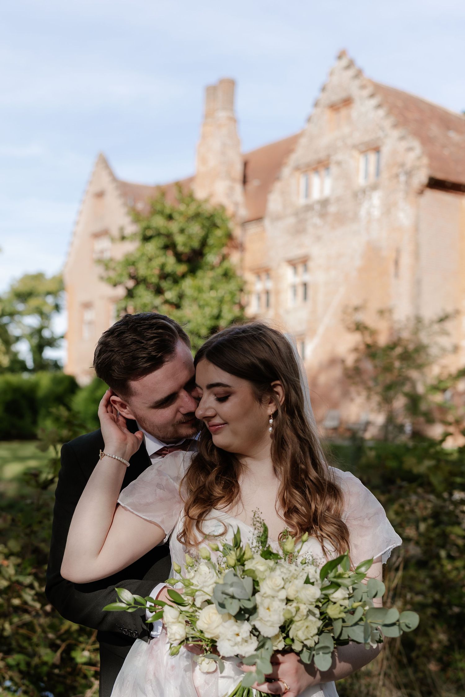 Groom tenderly embraces bride holding white bouquet with historic red brick manor in background.