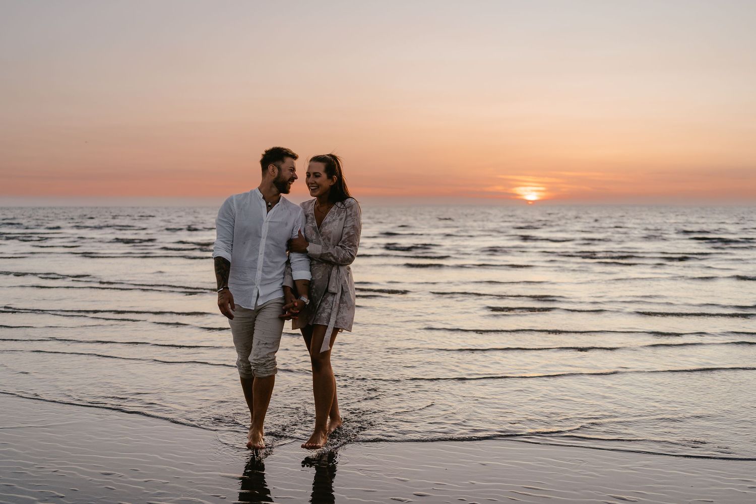 Romantic couple walking on wet beach at golden sunset, smiling and holding each other close.
