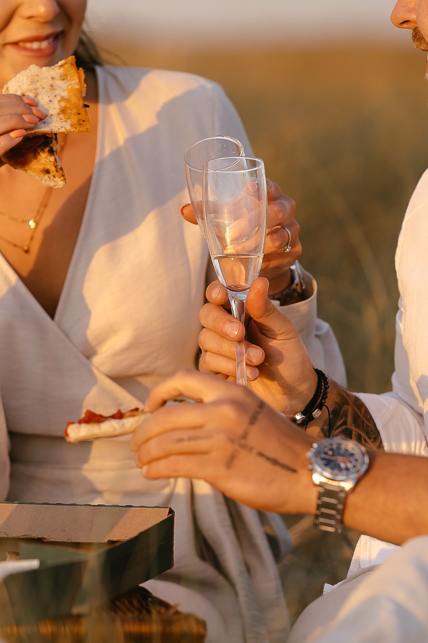 Close-up of couple clinking champagne flutes while eating pizza at a romantic outdoor picnic.