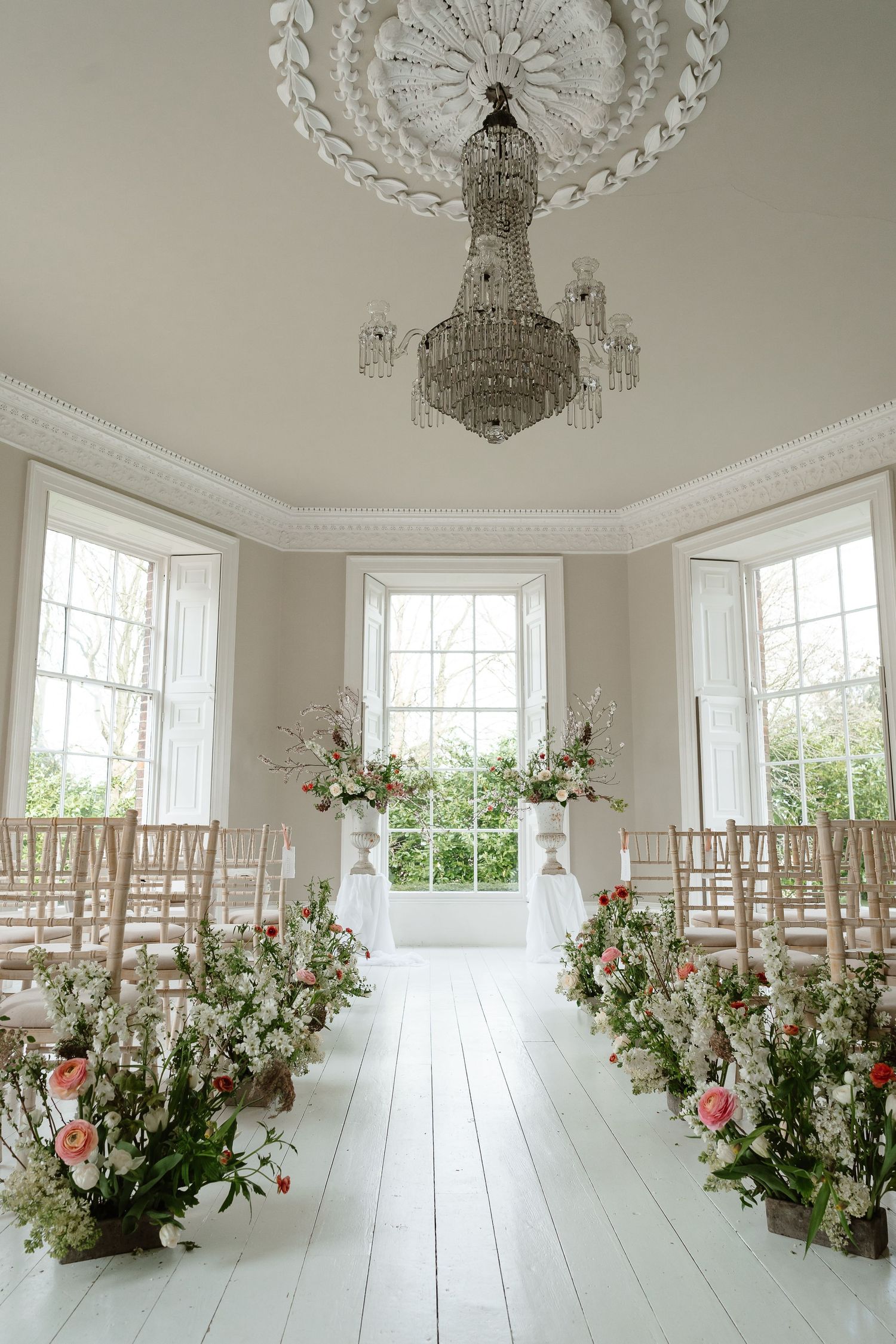 Elegant indoor wedding ceremony aisle lined with wildflower arrangements under crystal chandelier in bright white room.