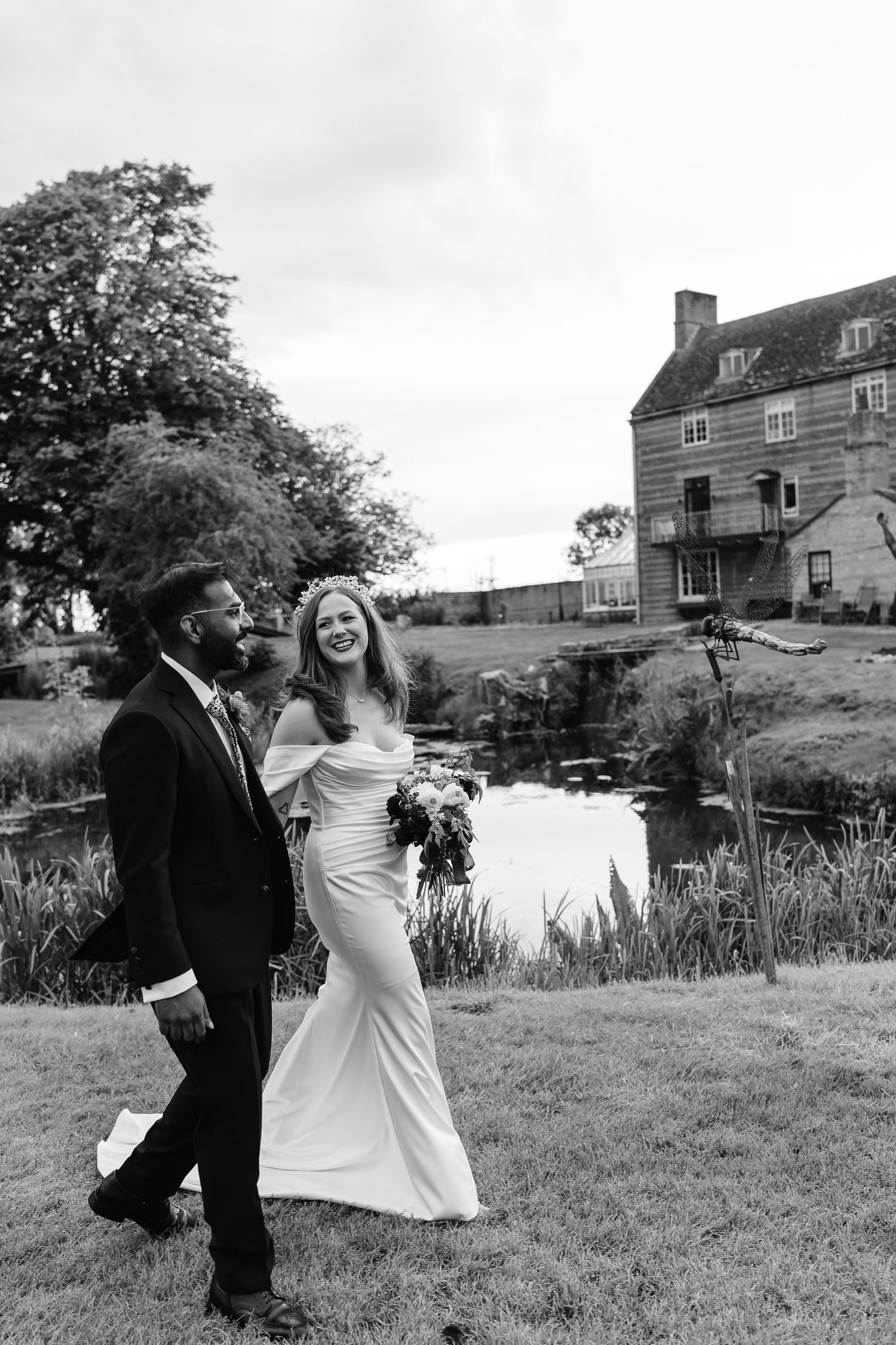 Smiling bride in off-shoulder gown and groom in black suit walking together at the beautiful Moleceys Mill wedding venue in Cambridgeshire