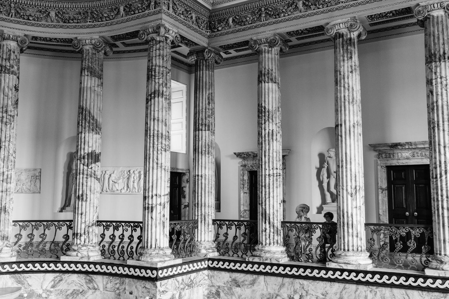 Black and white photo of grand neoclassical interior with tall marble columns, ornate railings, and classical sculptures.