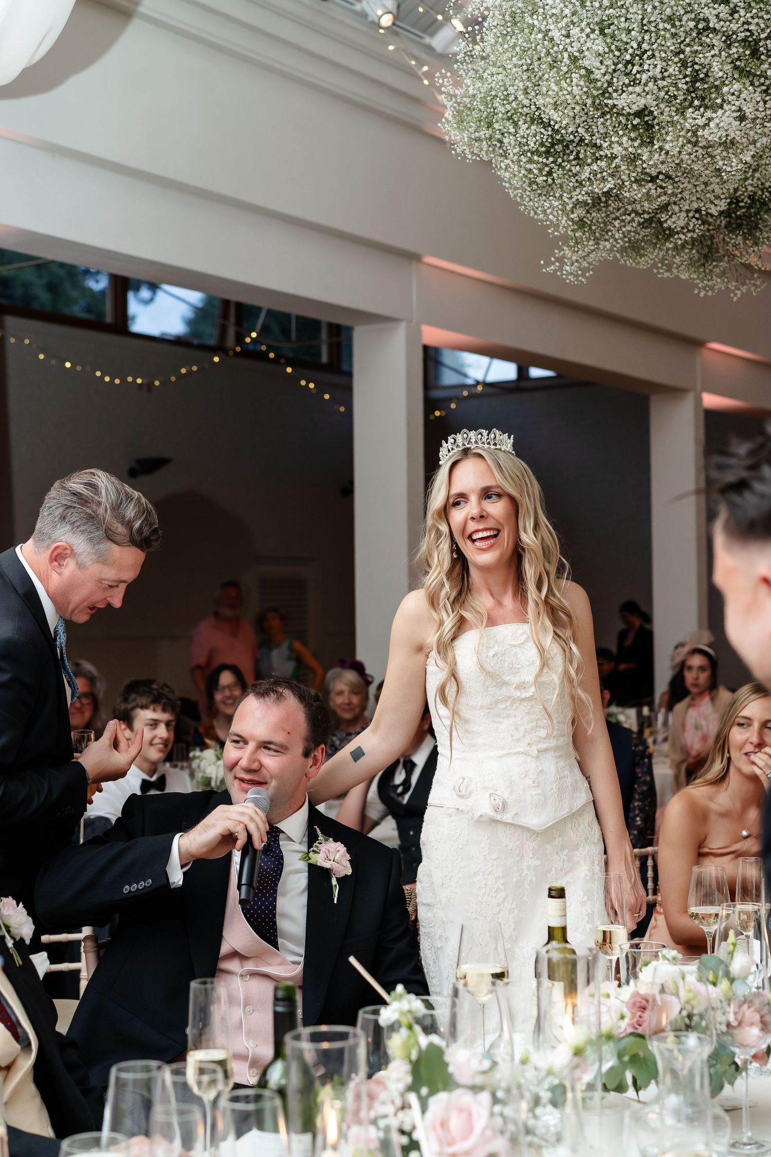 Radiant bride wearing tiara and white gown laughs during wedding reception speech at elegantly decorated table.