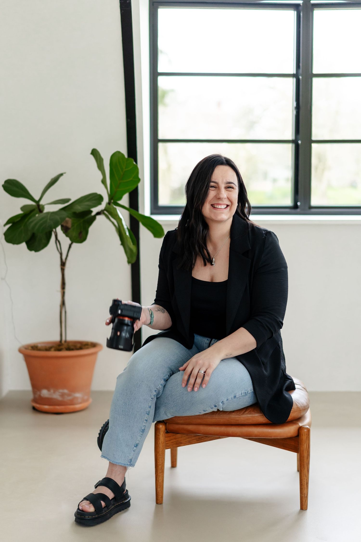 Smiling female photographer in black blazer and jeans sitting on wooden chair holding DSLR camera indoors.