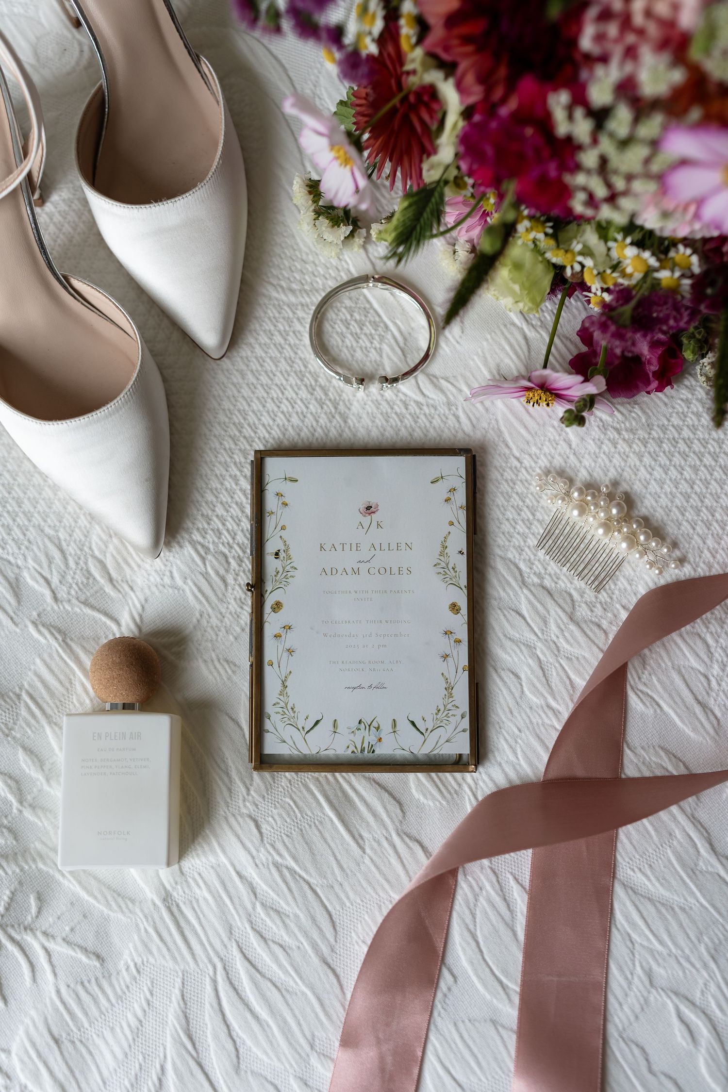 Elegant wedding flat lay with white heels, floral bouquet, gold ring, pearl hair comb, invitation, perfume, and pink ribbon.