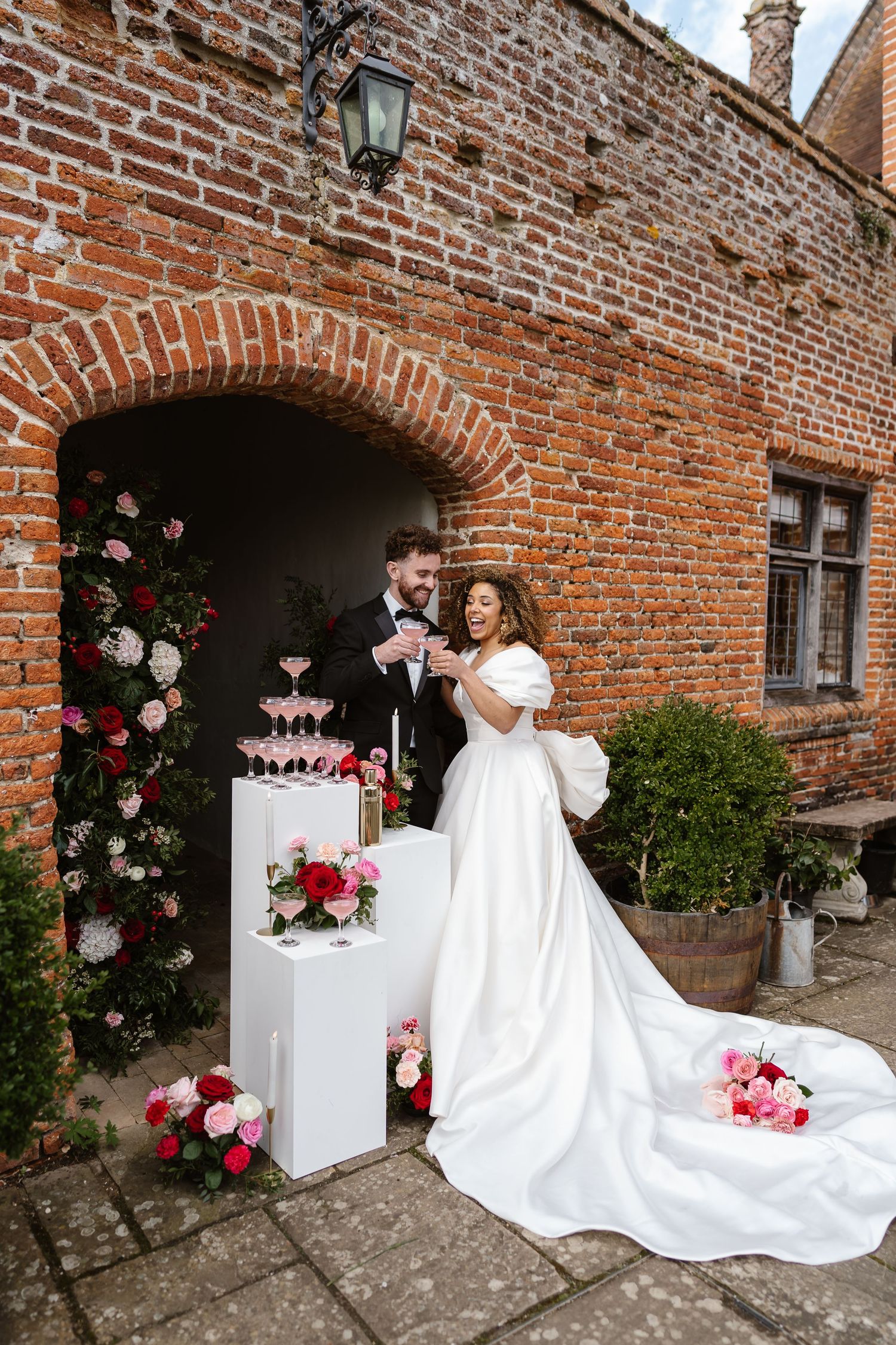 Bride and groom toast champagne tower beside floral arch at brick venue wedding.