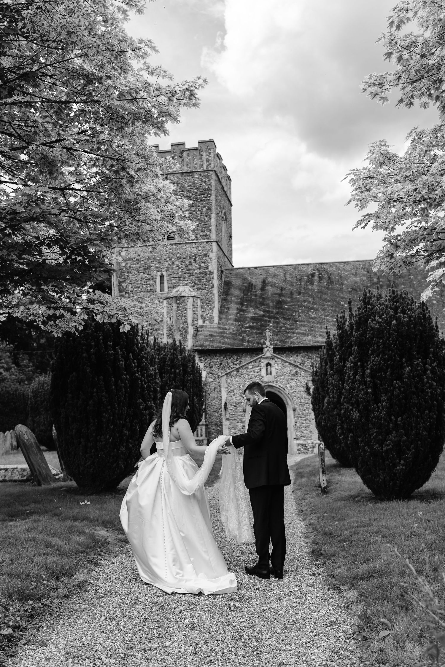 Black and white photo of groom spinning bride on gravel path outside rustic stone church with trees.