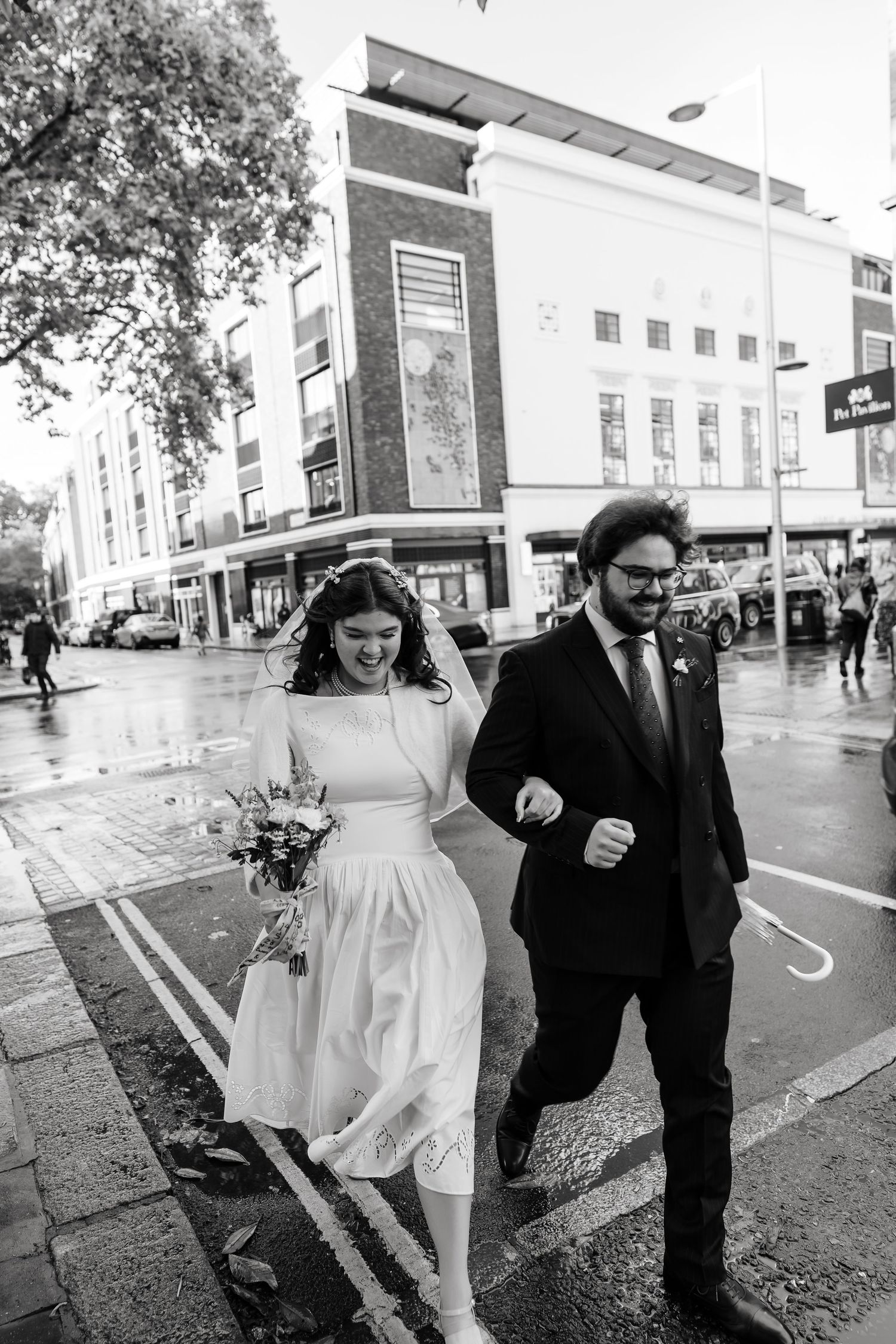 Black and white photo of bride and groom walking in the rain on a city street.