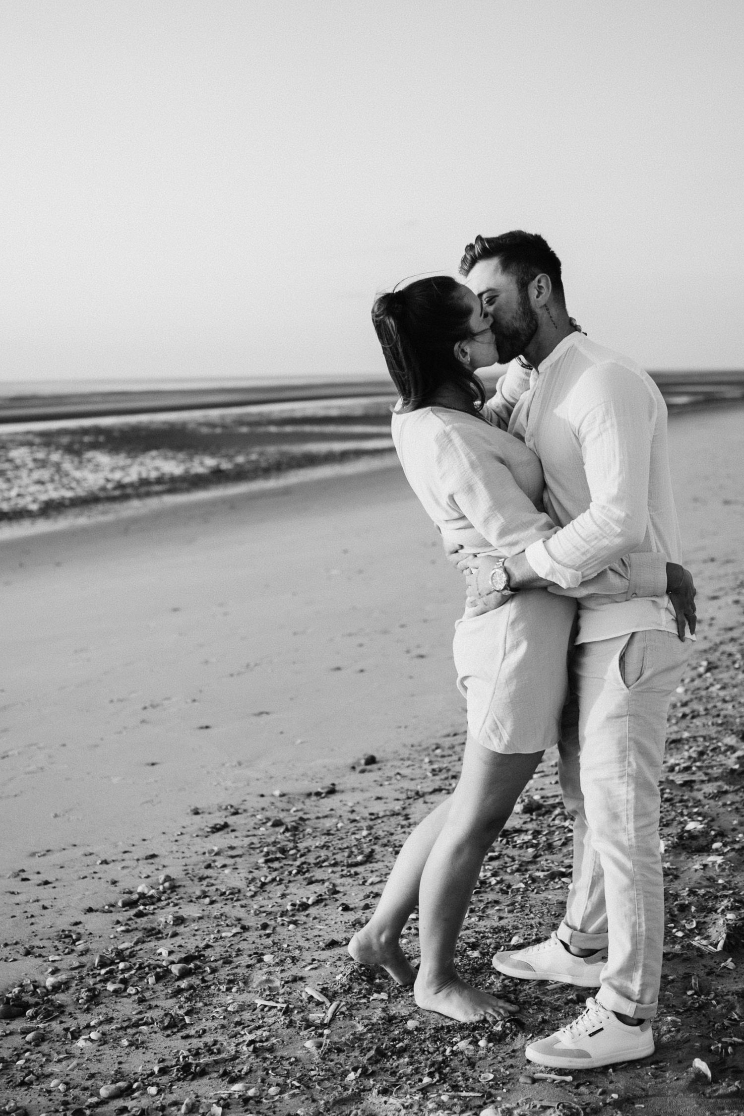 Black and white photo of couple kissing on beach with ocean in background.