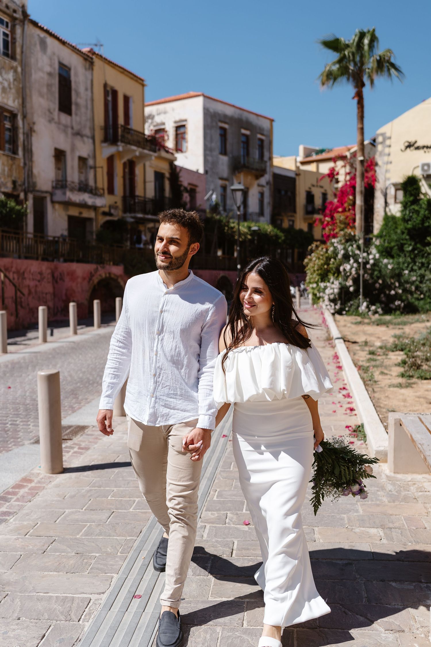 Bride carrying a bouquet and groom walking hand-in-hand through a sunny Mediterranean old town street.