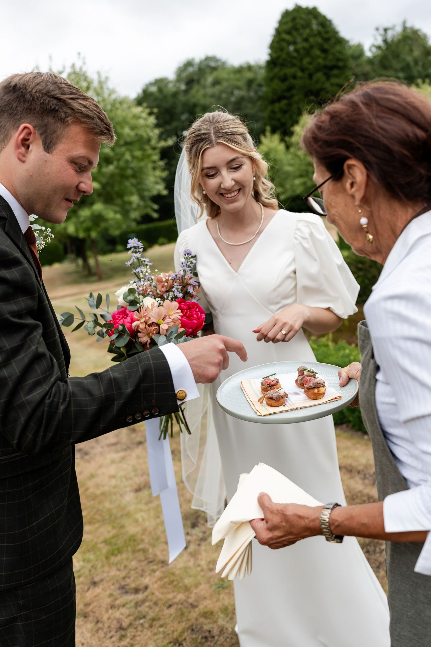 A smiling bride and groom sample canapes served by a caterer at their outdoor wedding reception.