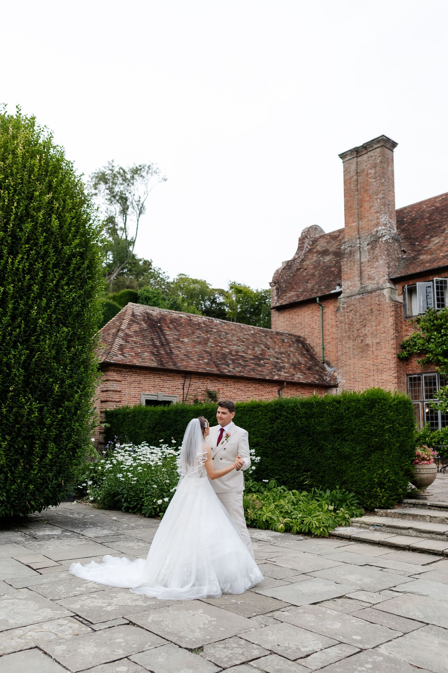 Bride and groom pose in garden of historic brick manor house on their wedding day.