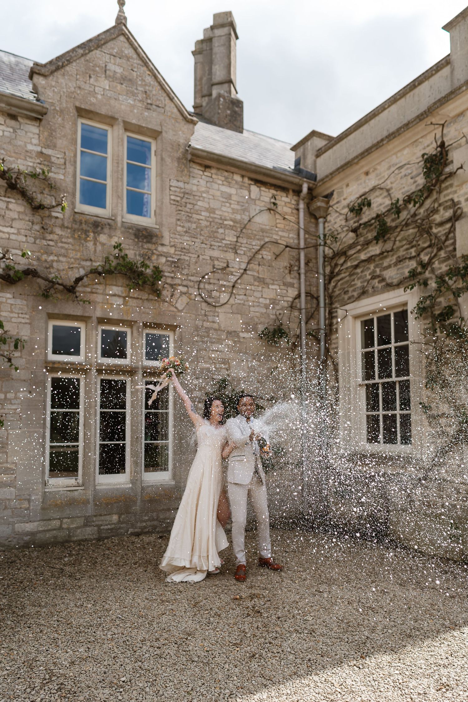 Joyful newlyweds celebrating with a champagne spray outside a beautiful historic stone manor house.