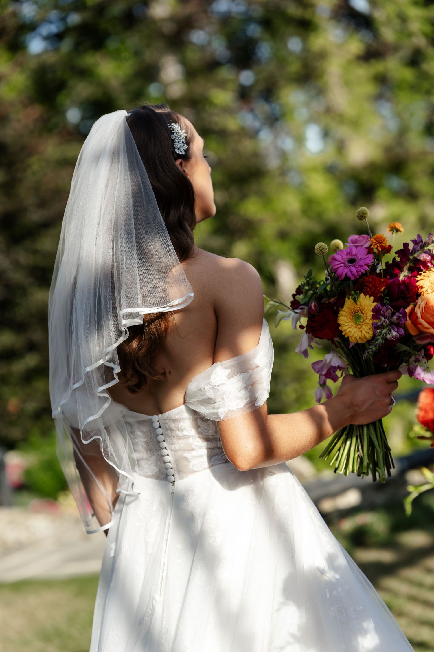 Bride in white gown and veil holds colorful bouquet with back to camera.
