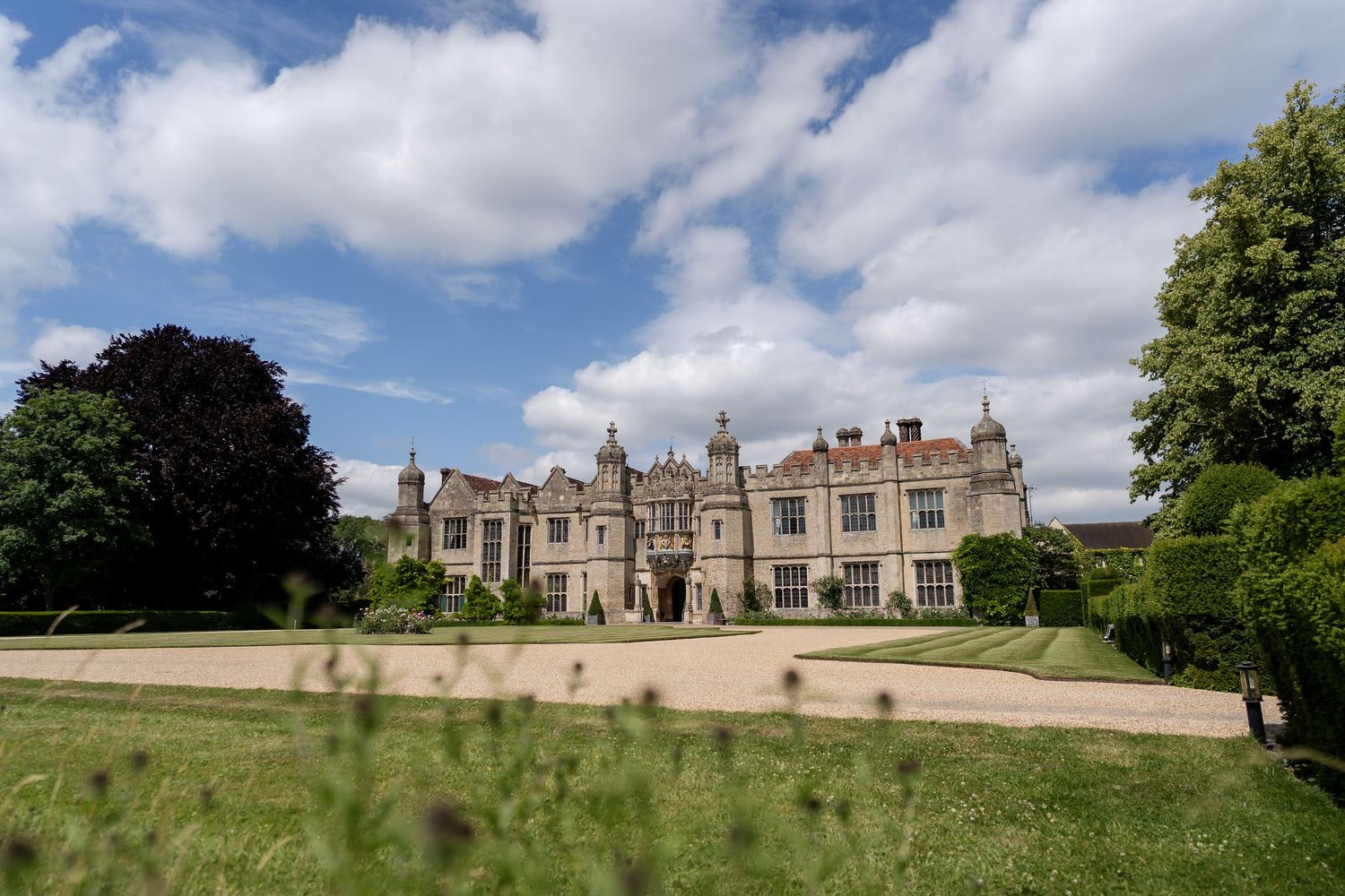 Grand historic English manor house with manicured grounds under dramatic cloudy blue sky.