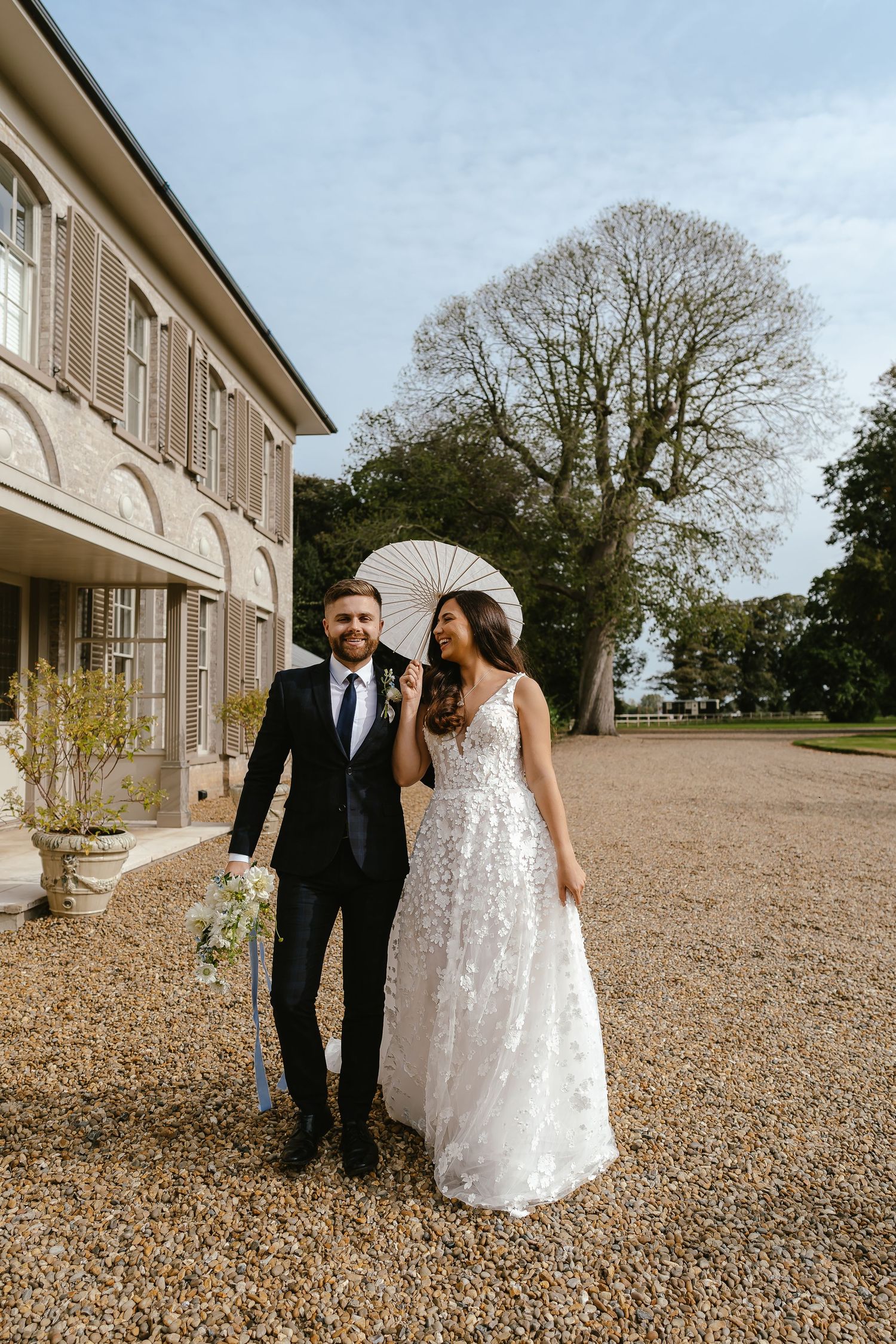 Bride and groom walking outside a grand estate, bride holding a white parasol, on a gravel path.
