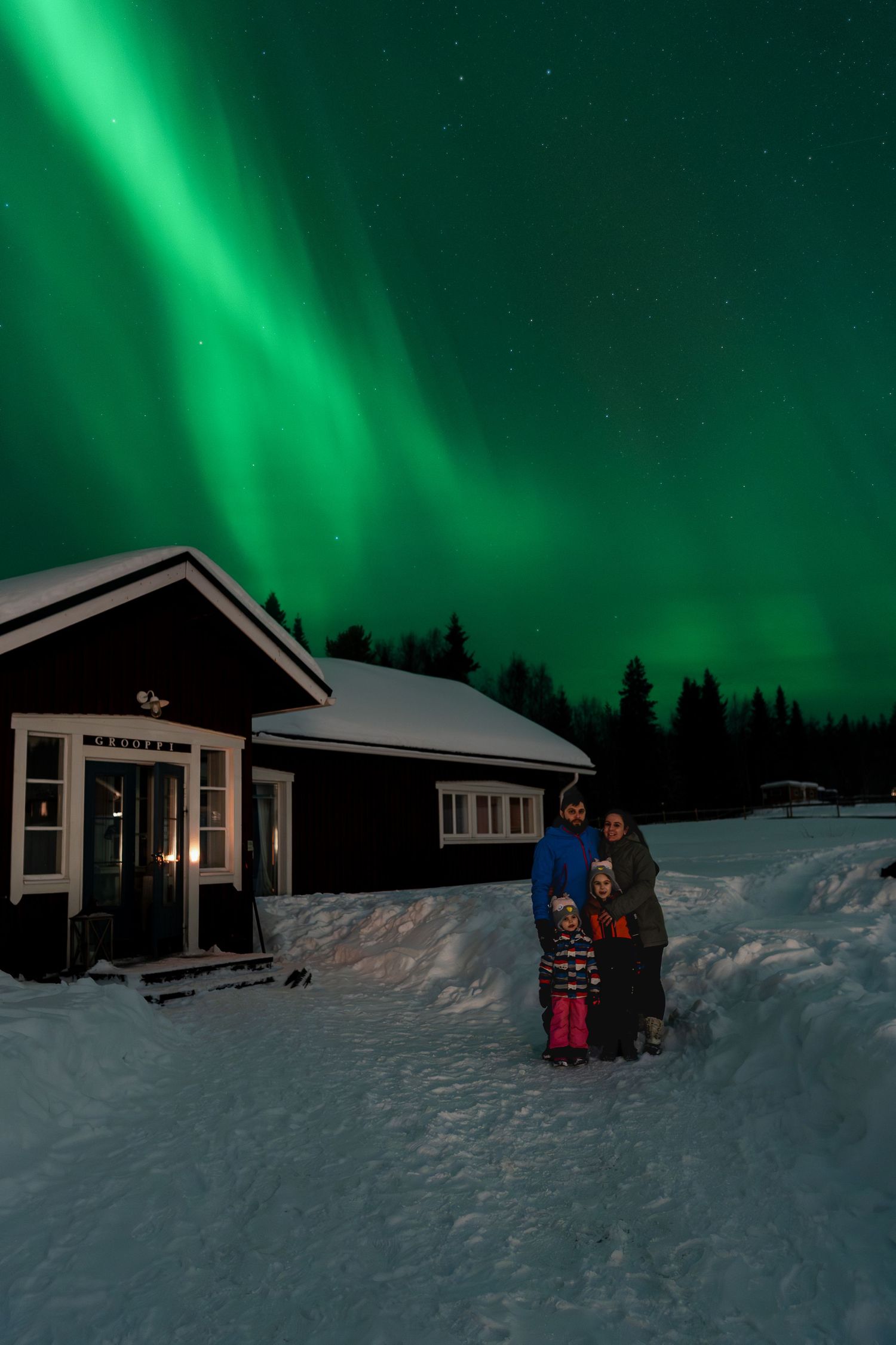 Family posing under vibrant green northern lights aurora borealis beside a snow-covered cabin in winter.