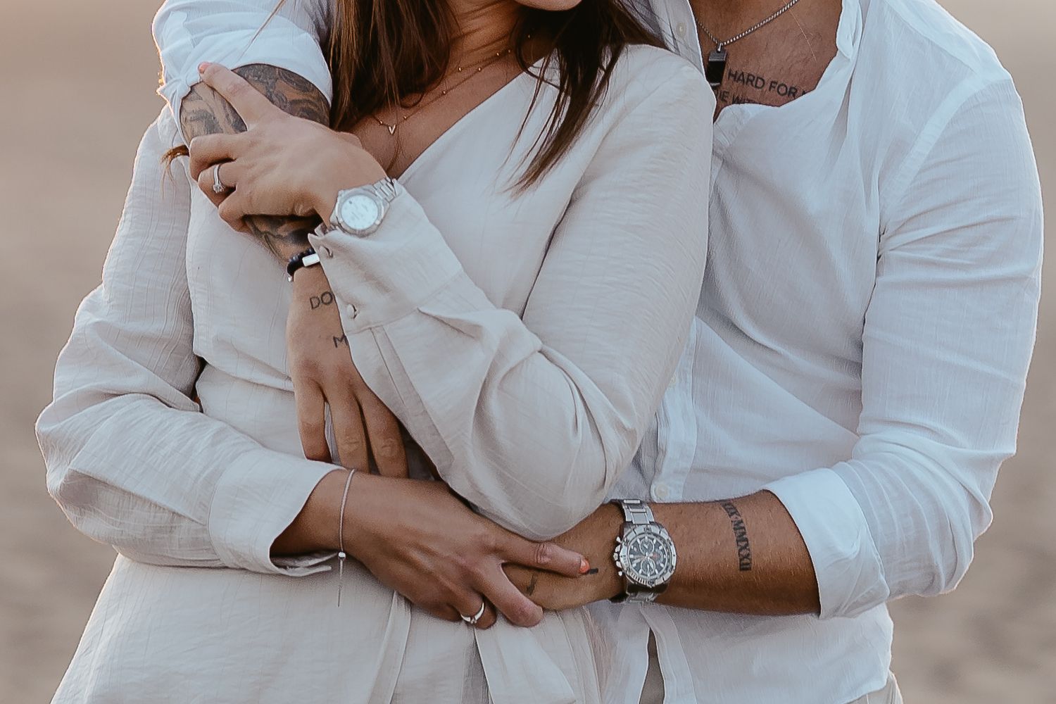 Close-up of a couple embracing, highlighting matching watches, tattoos, and neutral-toned outfits.