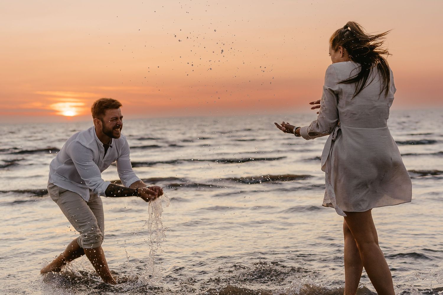 Man splashing water at laughing woman on beach during warm evening sunset.