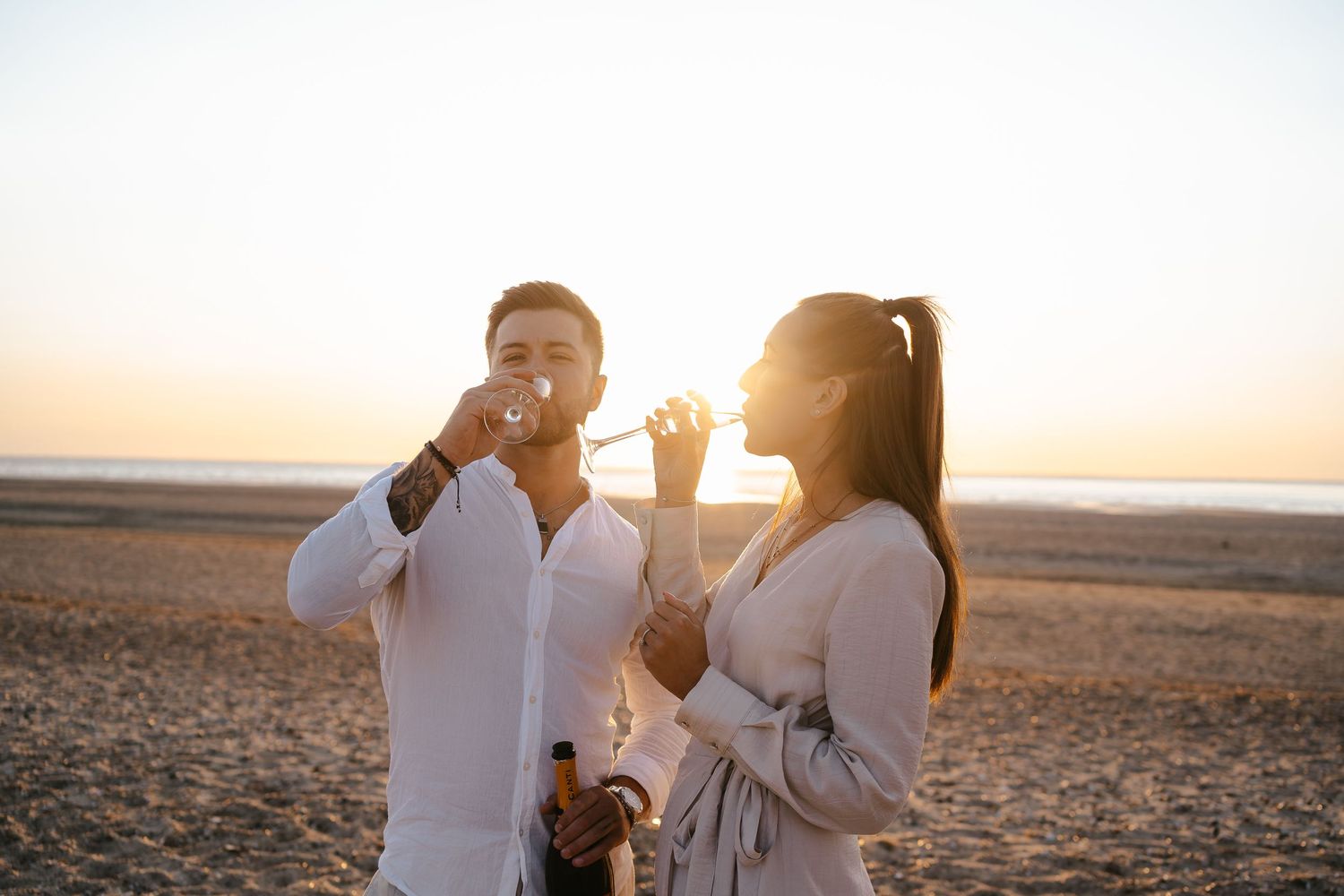 Happy couple drinking champagne together on a beach during a beautiful golden hour sunset.