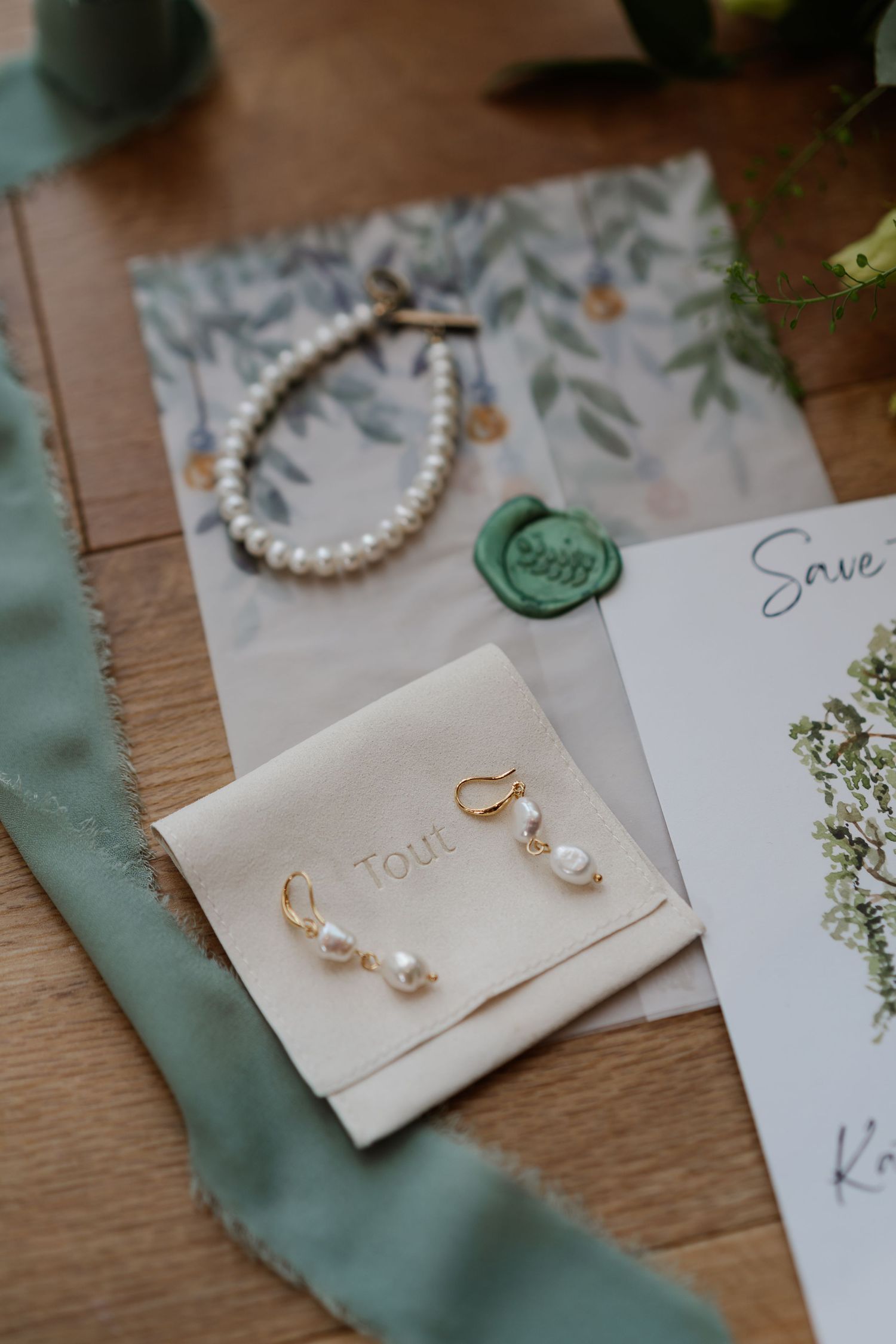 Elegant wedding flat lay featuring pearl earrings, bracelet, green wax seal stationery, and sage ribbon on wood.