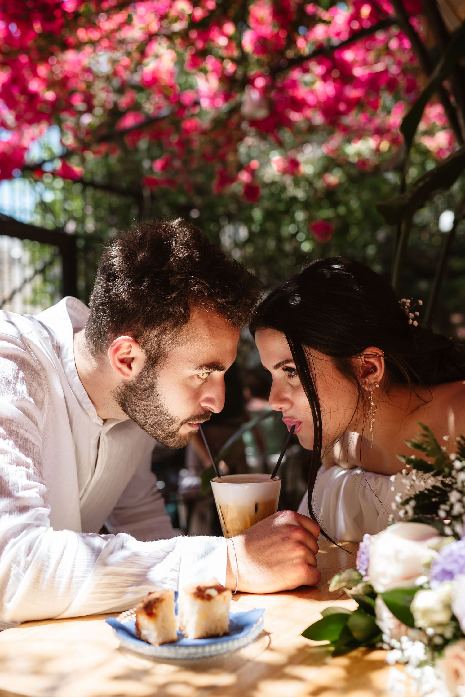Bride and groom leaning close together sharing an iced coffee under vibrant pink bougainvillea blooms.