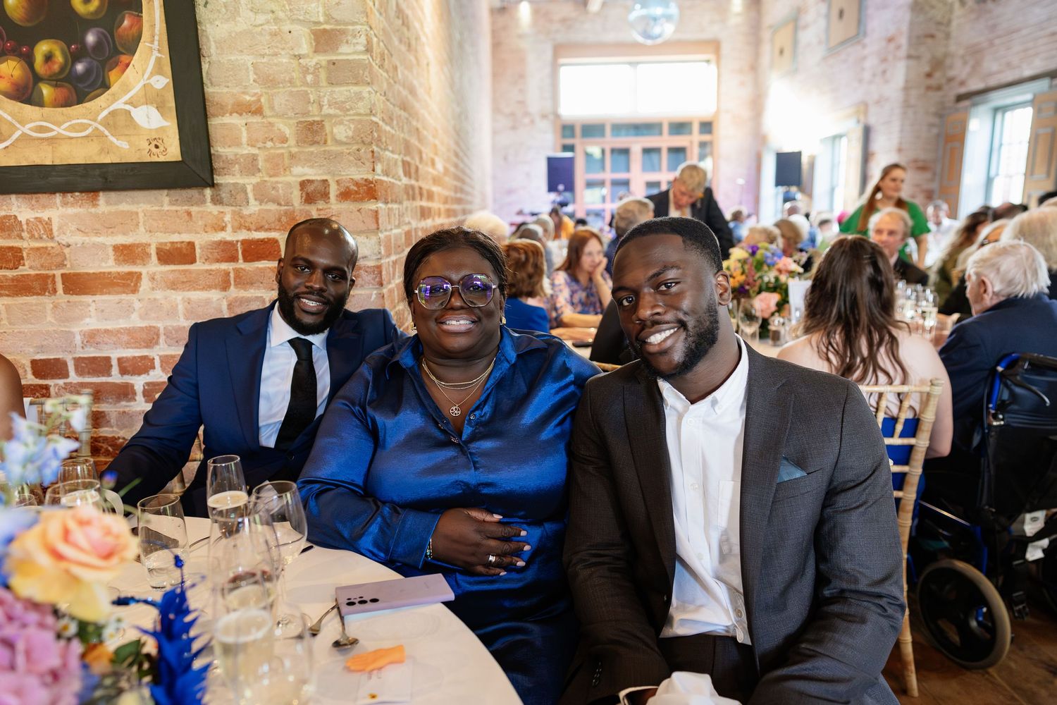 Three smiling wedding guests pose together at a reception table in a warmly lit rustic venue.