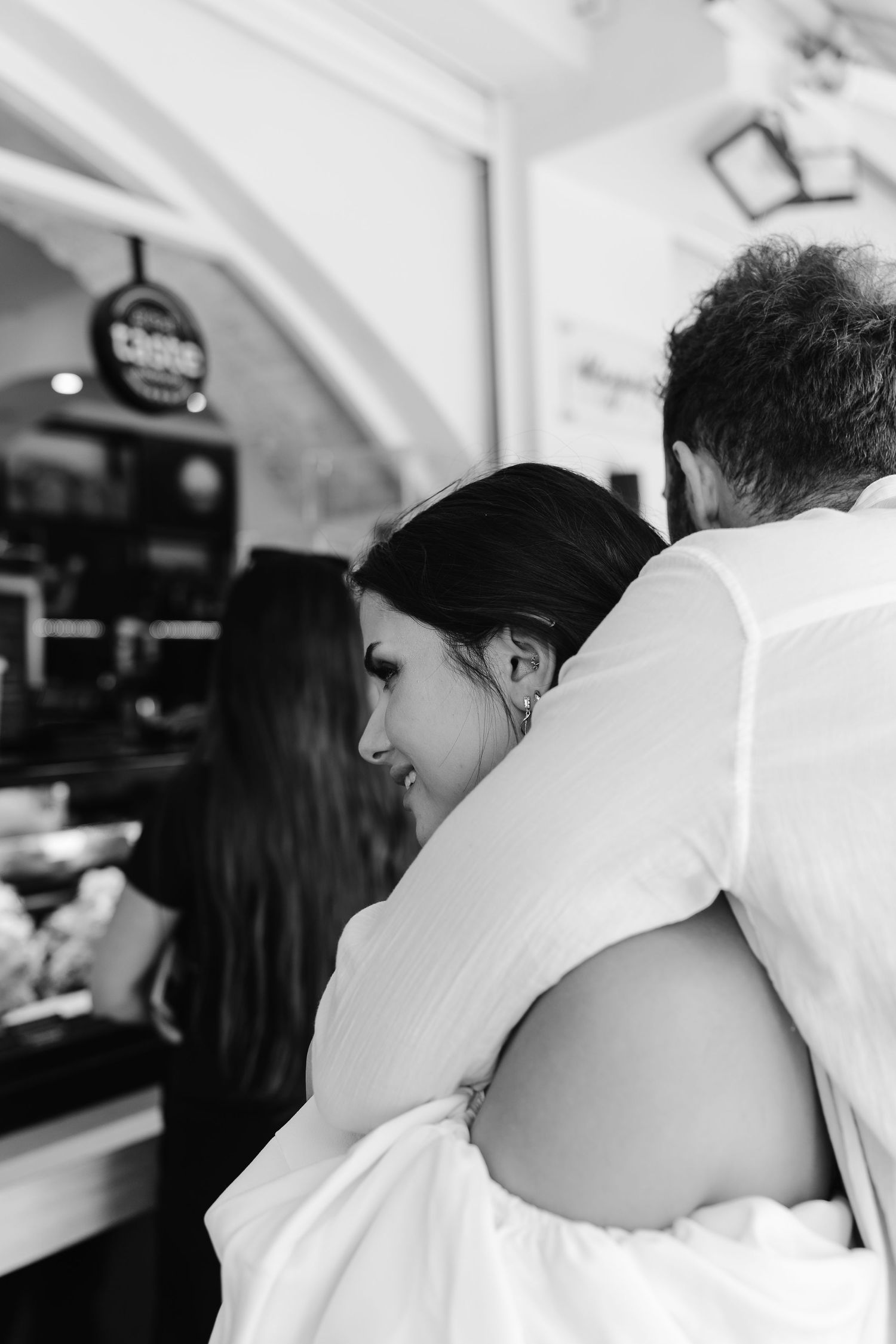 Black and white photo of bride embracing groom from behind inside a cozy cafe setting.