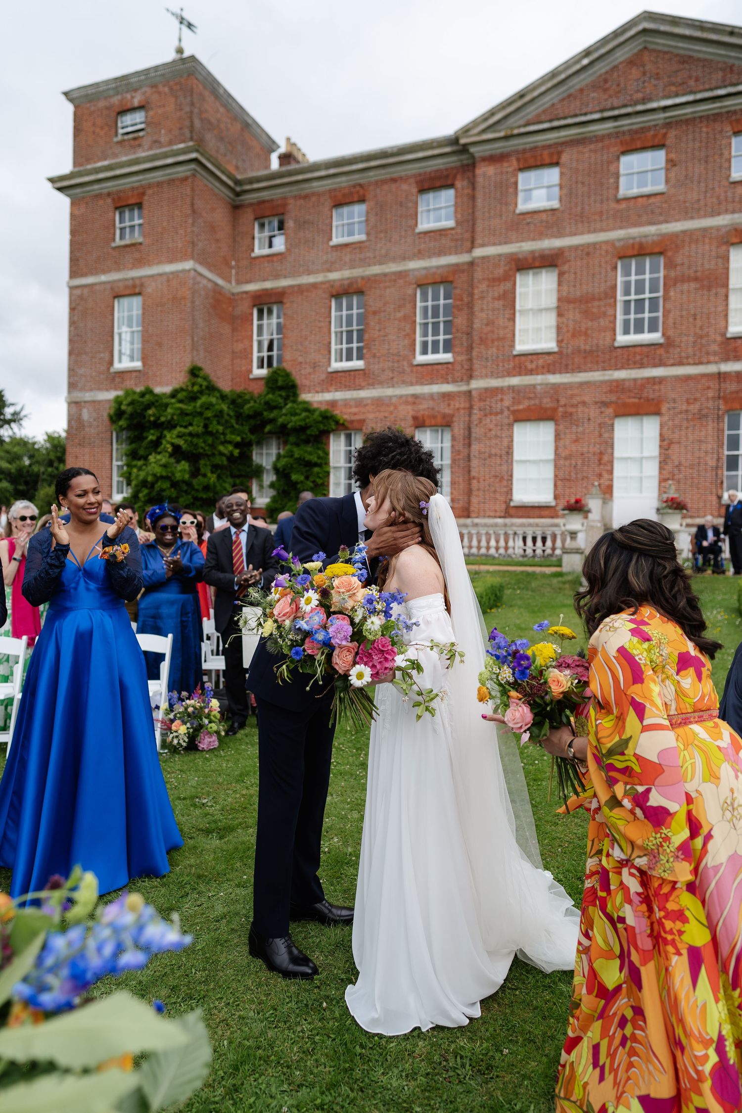 Bride and groom sharing first kiss at outdoor wedding ceremony on lawn of grand red brick estate.
