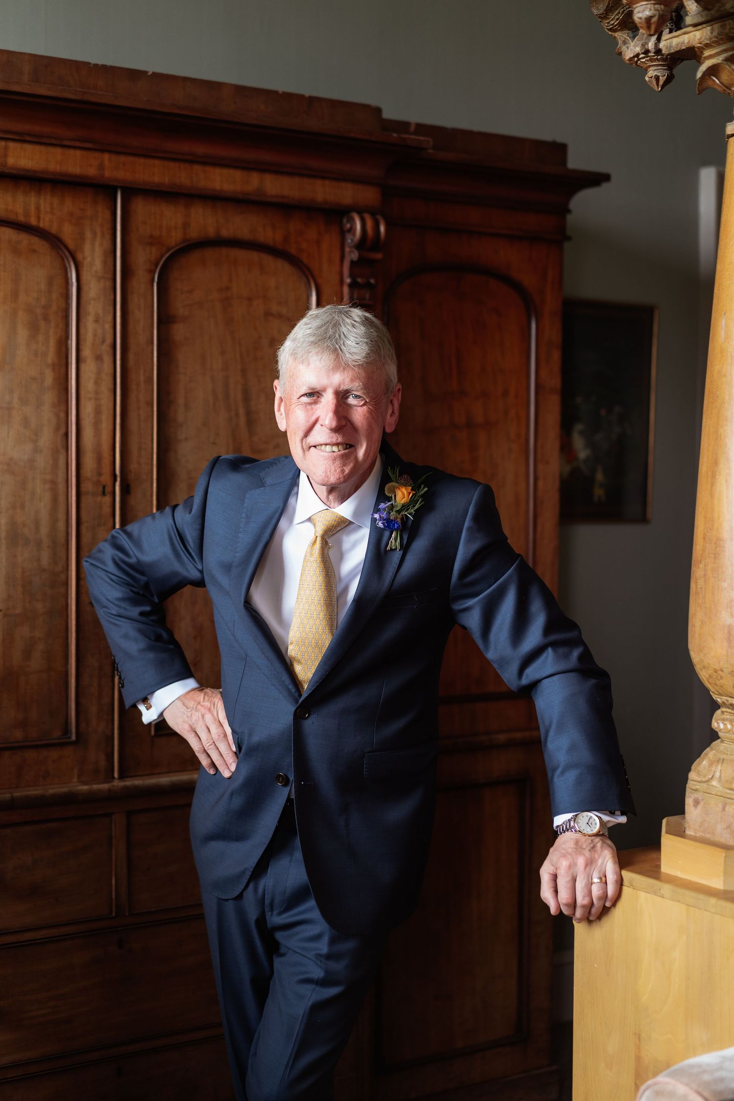 Elegant older gentleman in navy suit with gold tie and floral boutonniere leaning against wooden armoire.