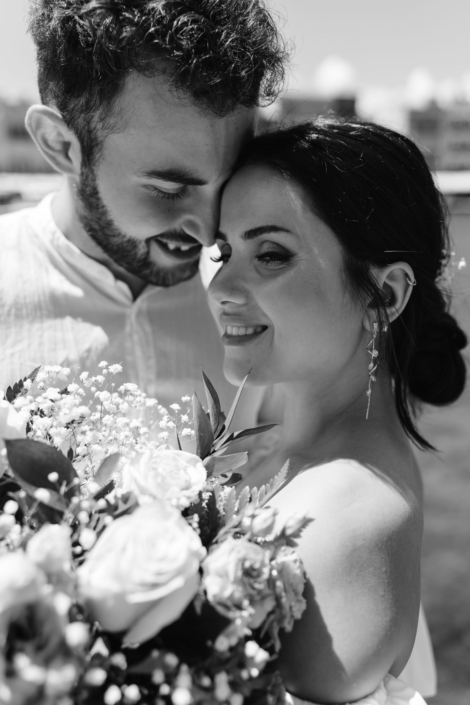 Intimate black and white portrait of a smiling bride and groom touching foreheads, bride holding a rose bouquet.