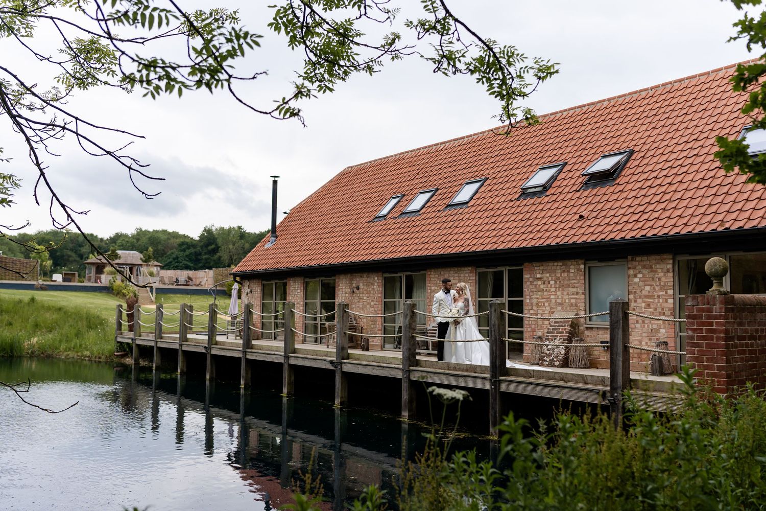 Bride and groom posing on a wooden deck over water beside a charming brick barn wedding venue.