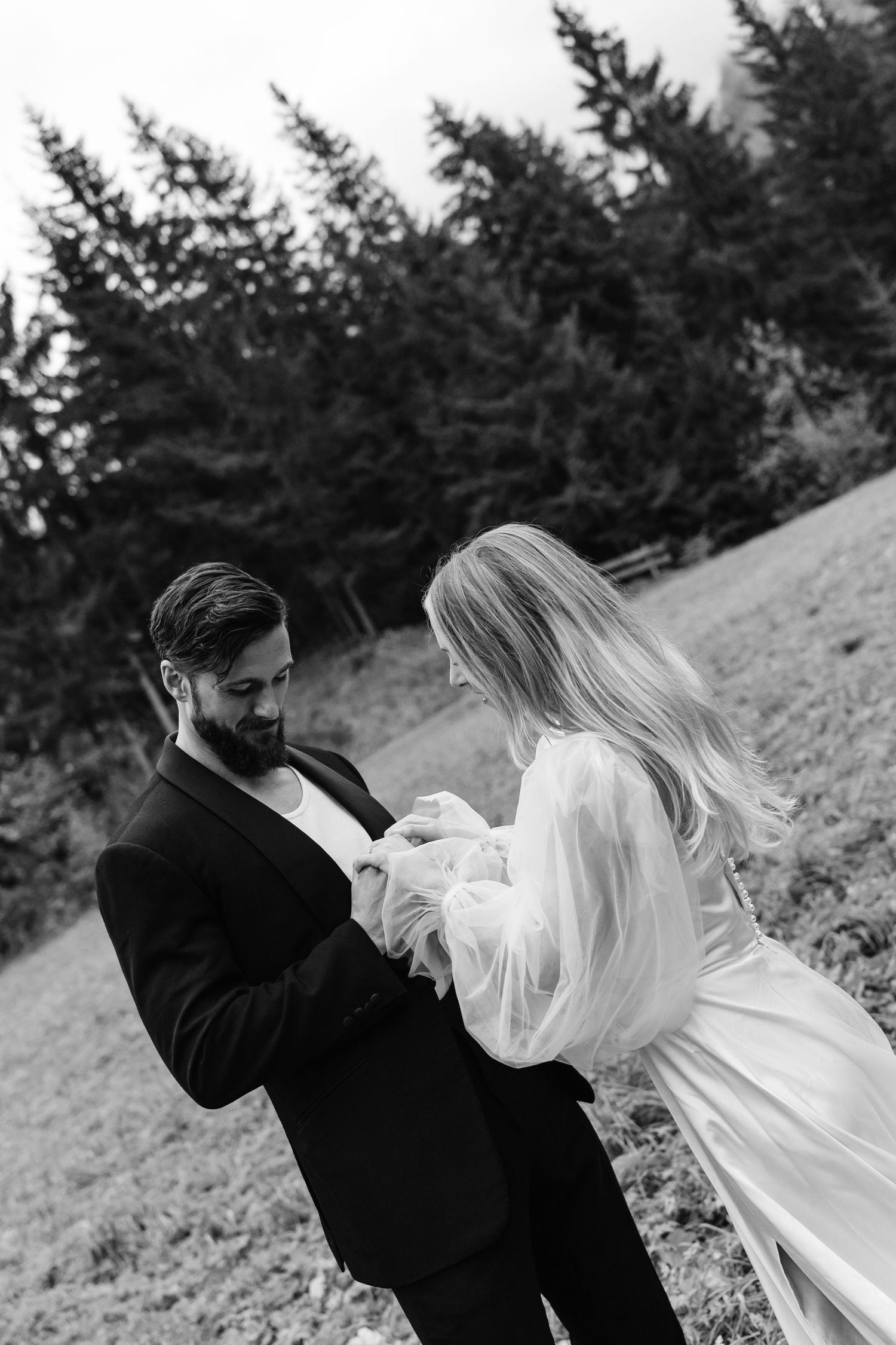 Black and white wedding photo of bride placing ring on groom's finger outdoors with evergreen trees in background.