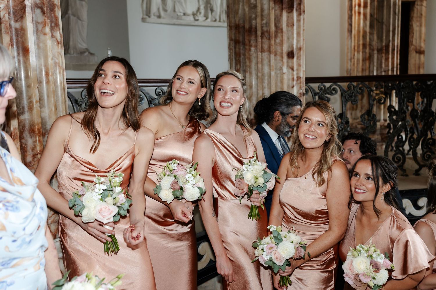 Bridesmaids in blush satin dresses holding white floral bouquets laughing joyfully at an elegant wedding ceremony.