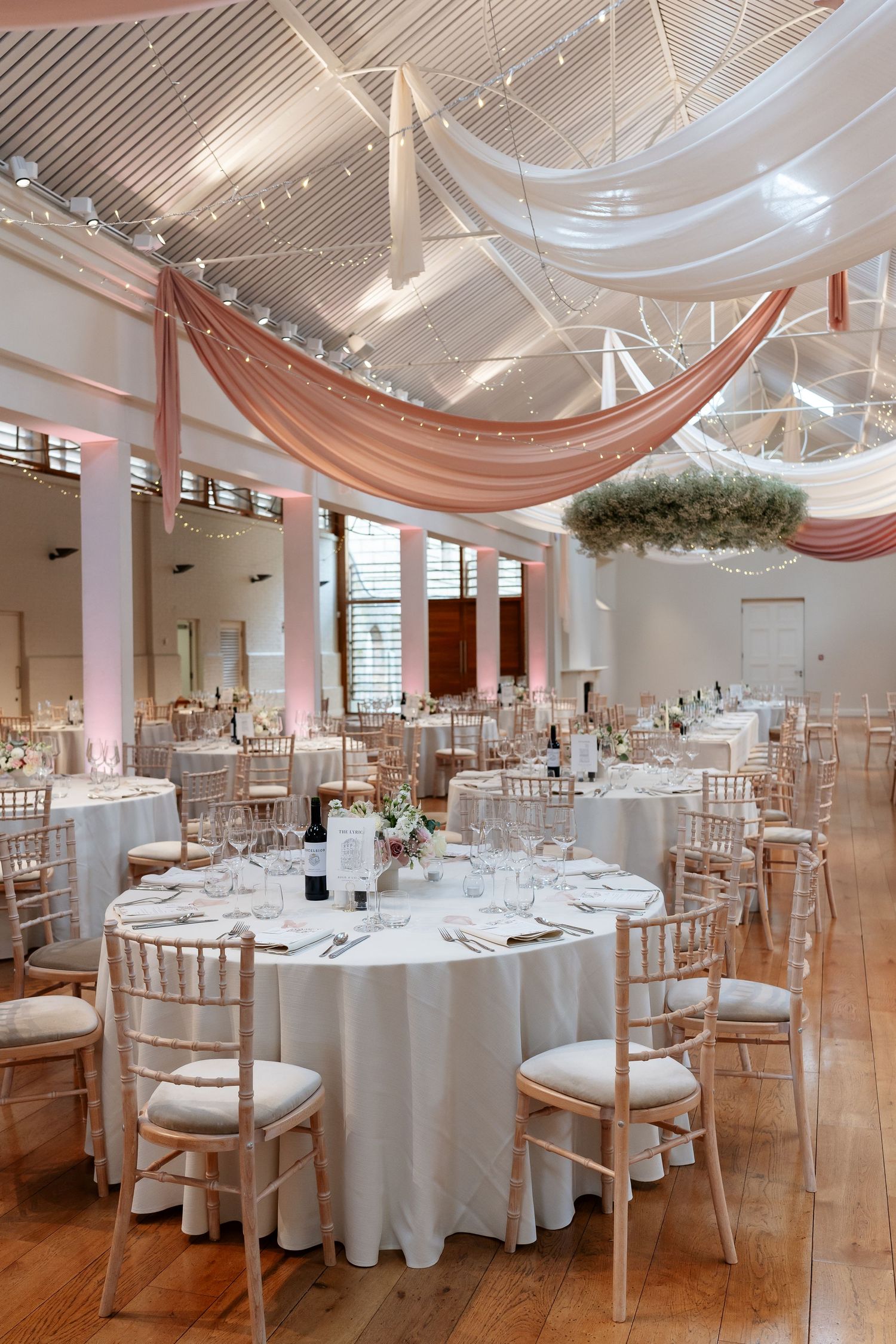 Elegant wedding reception hall with round tables, chiavari chairs, white linens, and pink draped ceiling decor.