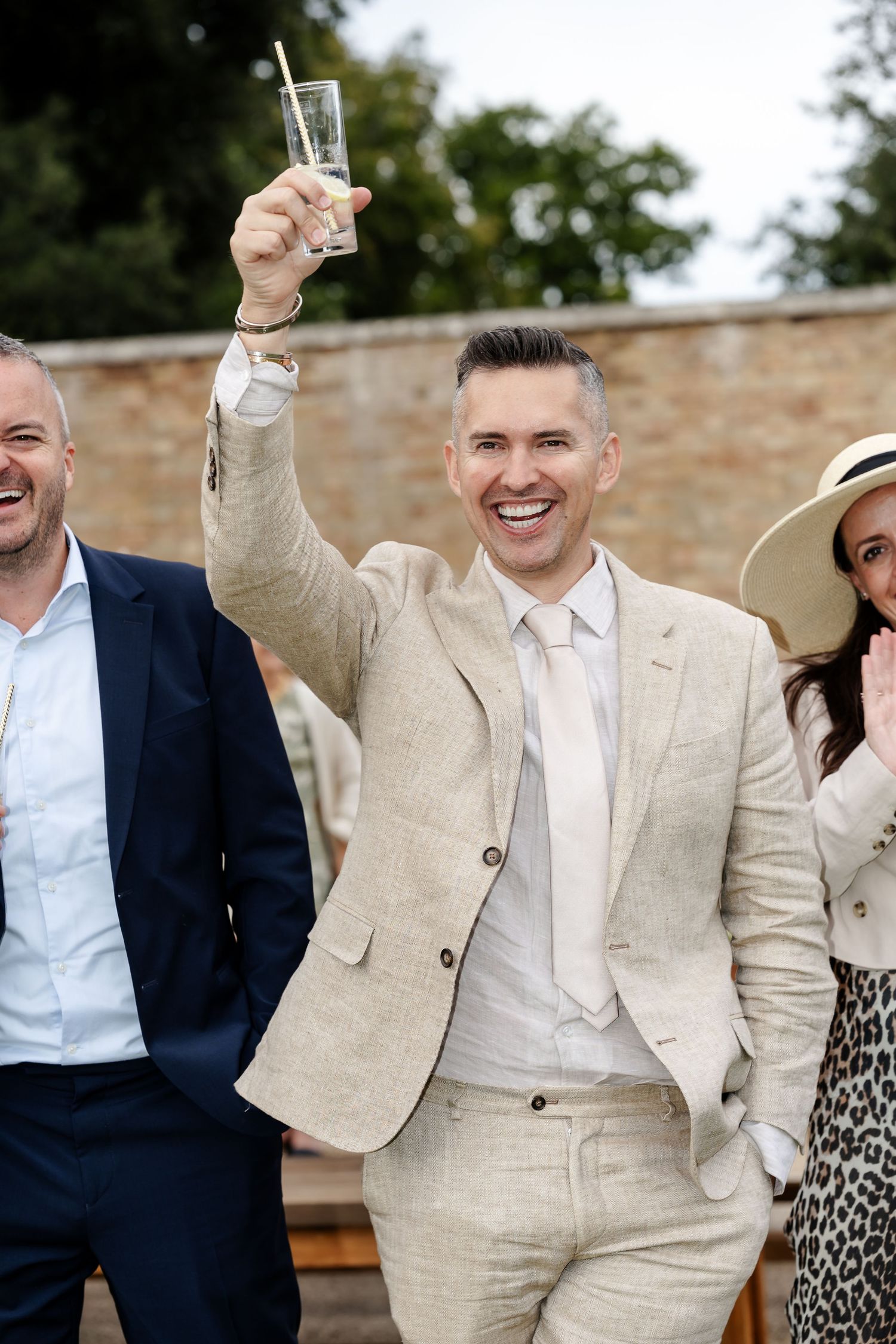 Cheerful wedding guest in beige suit raising glass in toast at outdoor wedding celebration.
