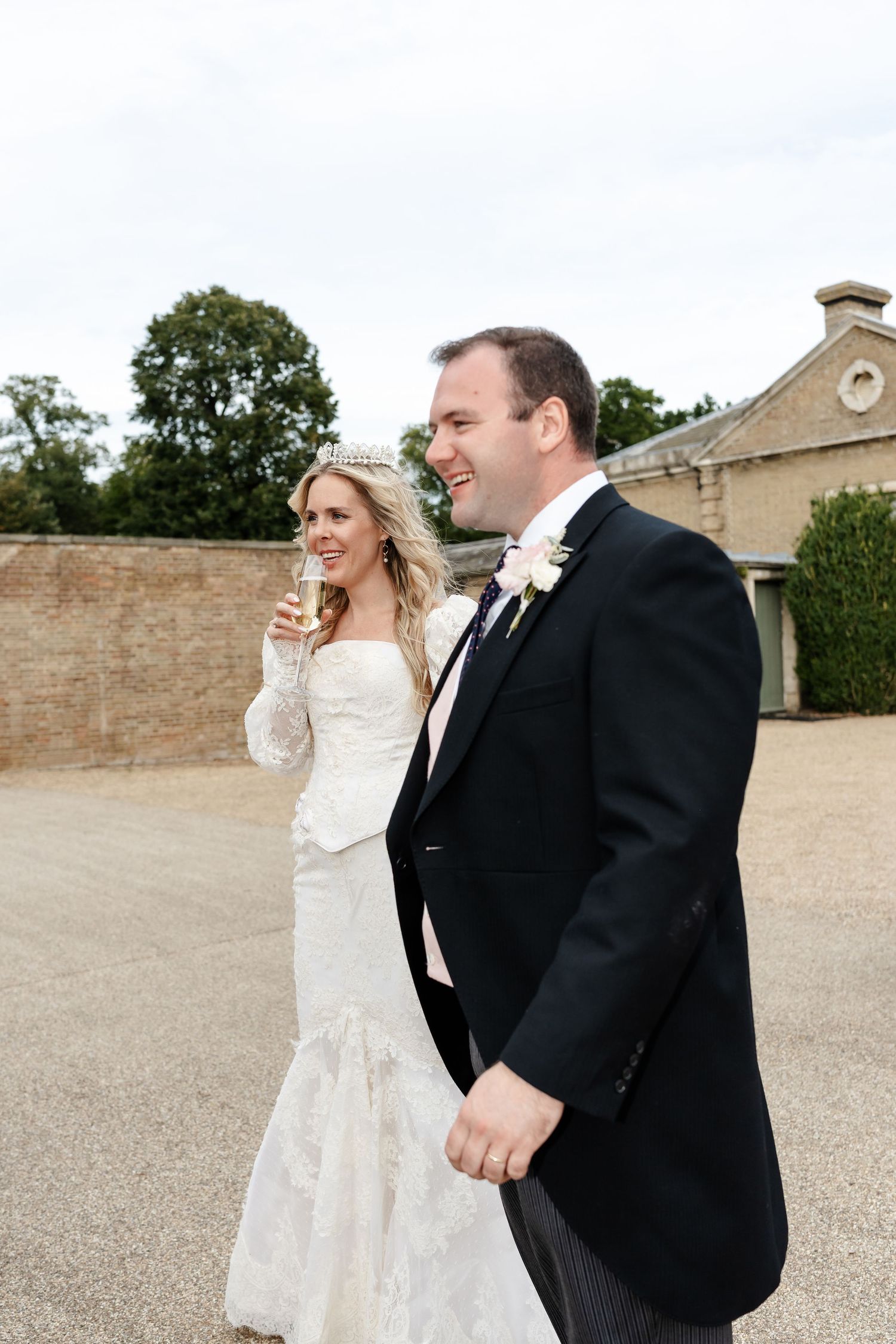 A smiling bride in a lace gown and tiara holds champagne beside a laughing groom in a dark suit at an outdoor wedding.