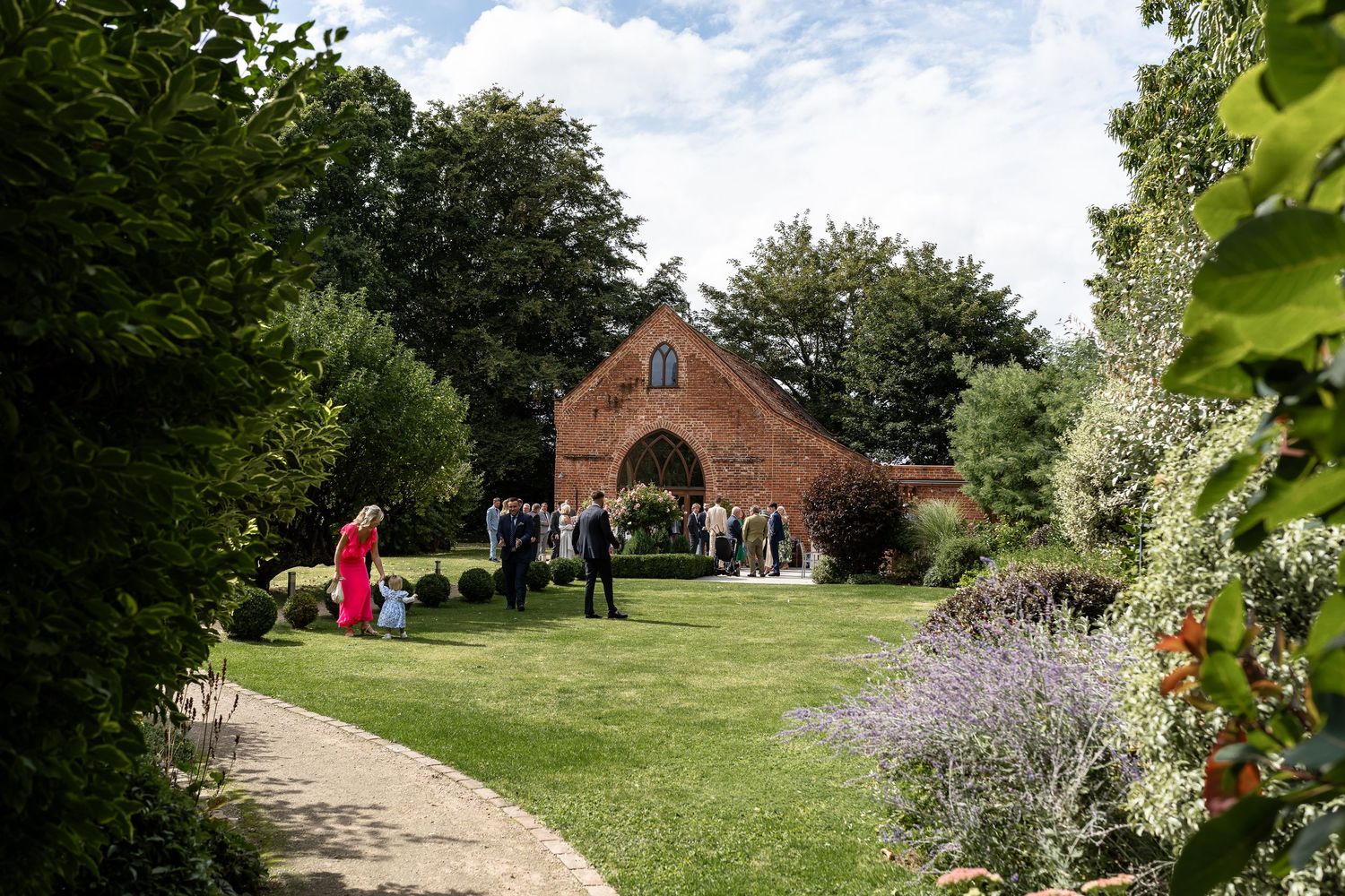 Guests gather on a lush green lawn beside a charming red brick chapel surrounded by beautiful gardens.