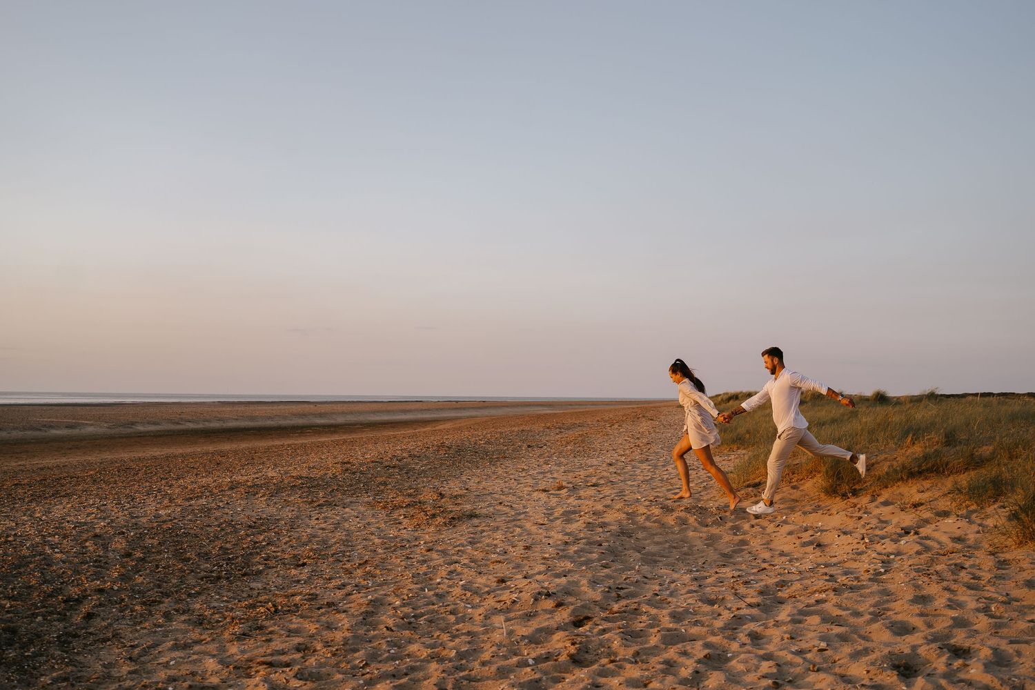 Joyful couple in white outfits holding hands and running along a vast sandy beach at dusk.