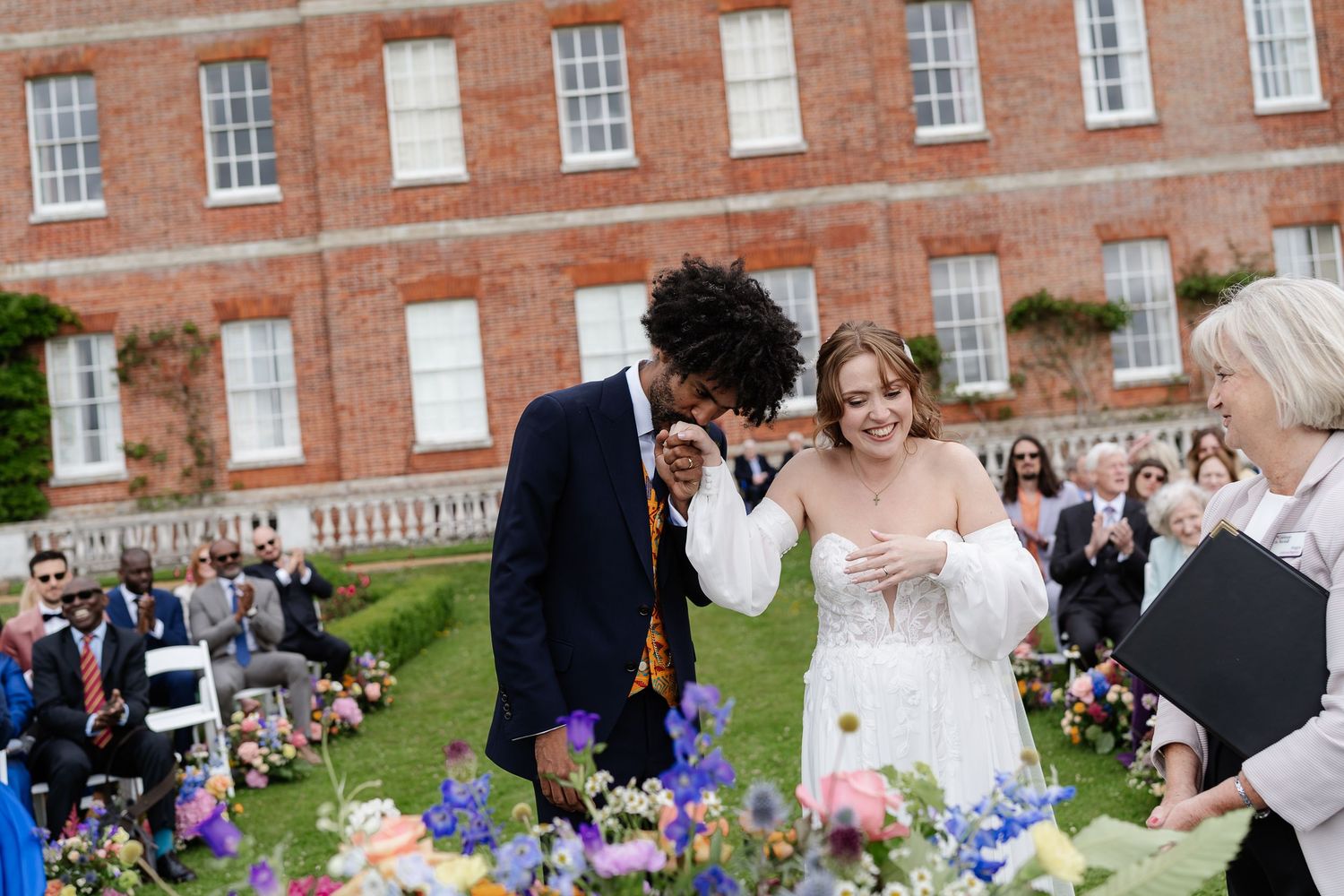Joyful newlyweds laugh together after ceremony in front of elegant red brick manor with colorful flowers.