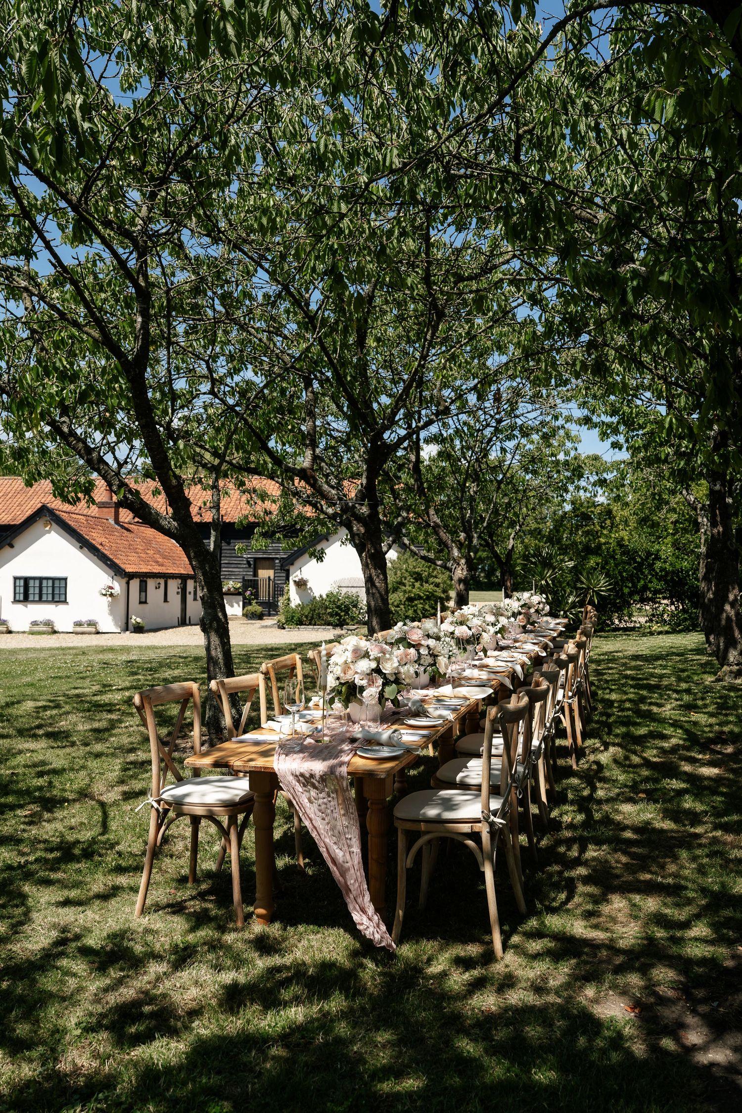 Long rustic wooden wedding reception table set outdoors under green trees in sunny garden.
