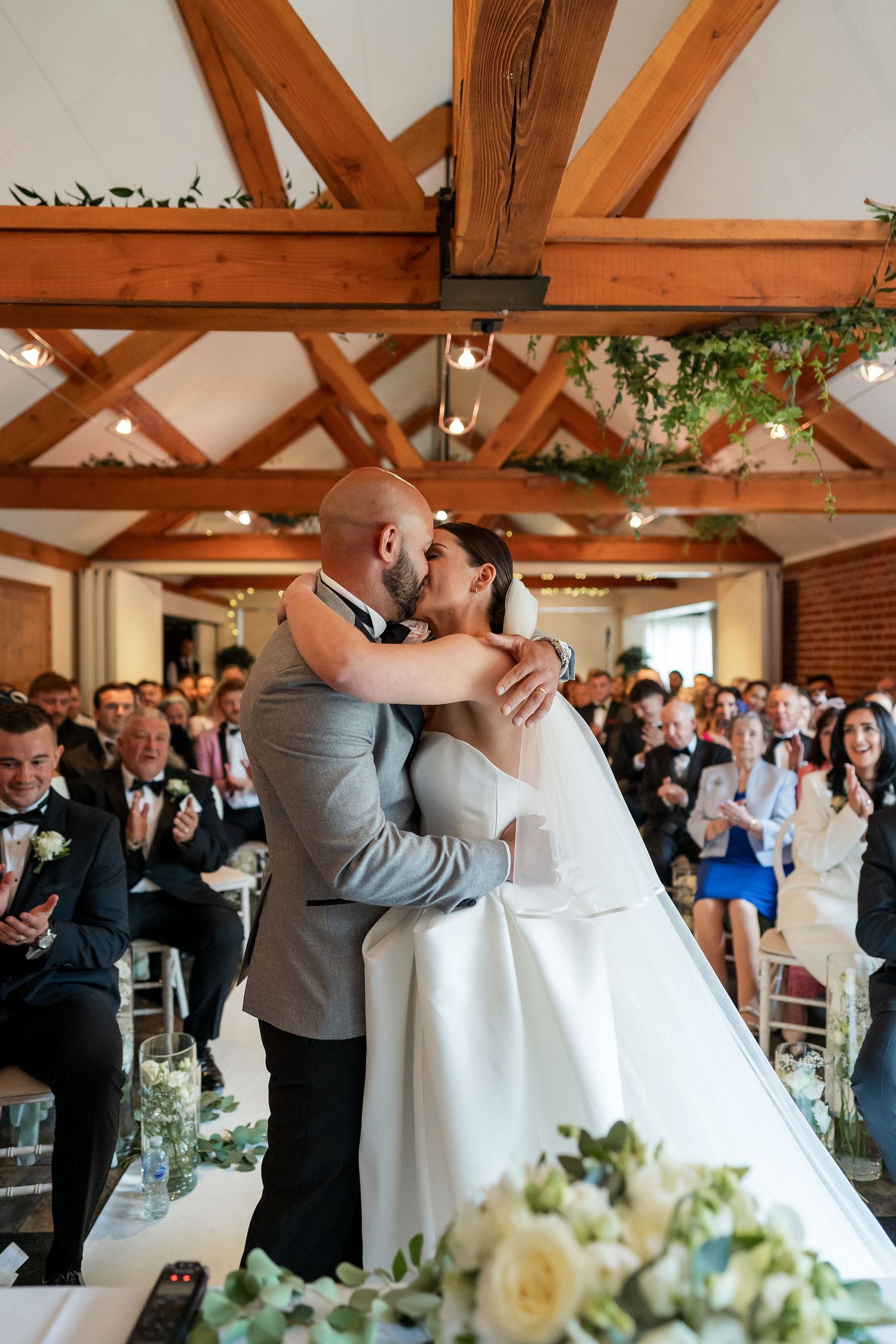 Bride and groom share first kiss at wedding altar surrounded by applauding guests.