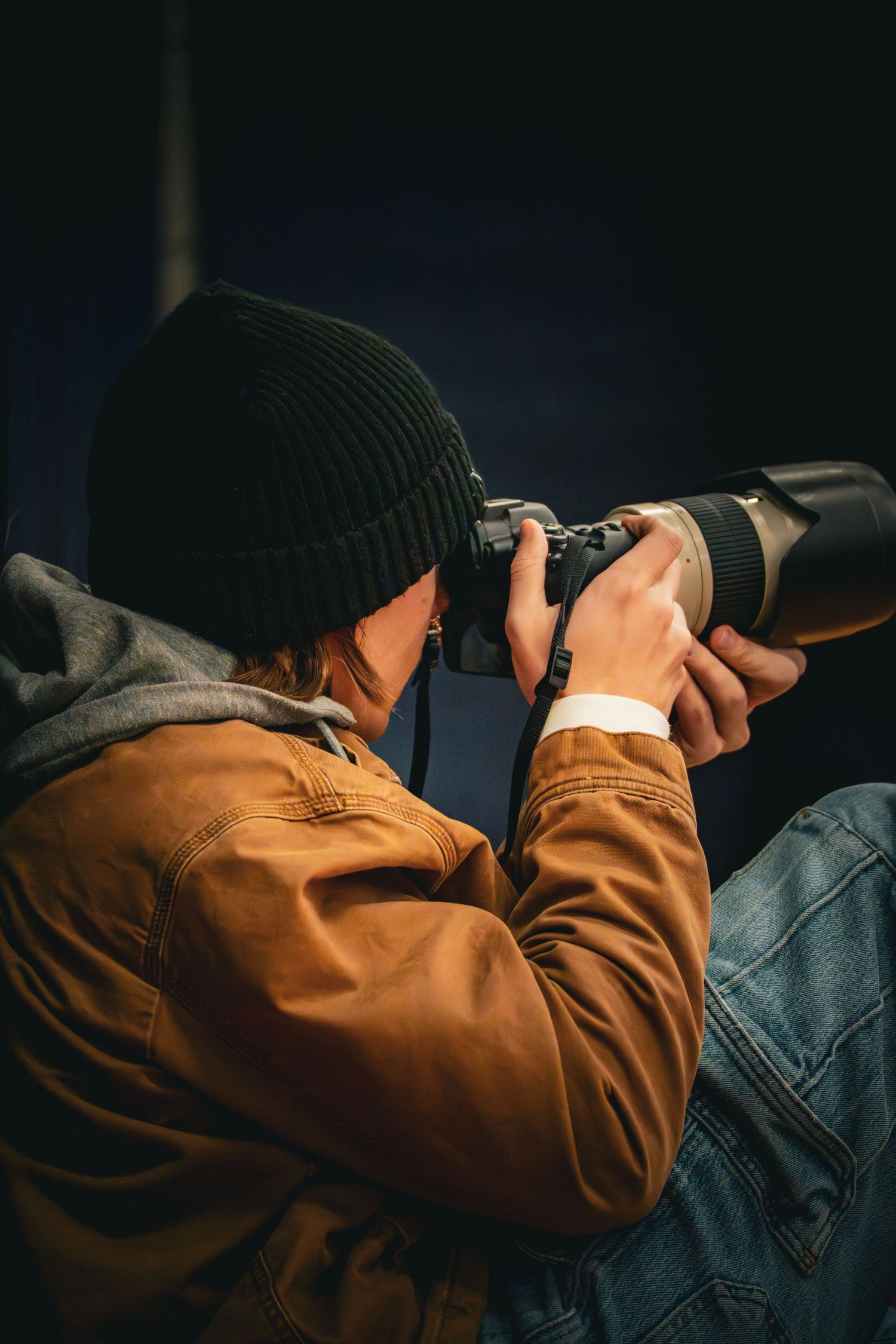 Photographer in brown jacket and black beanie captures shot with telephoto lens camera in dark studio setting.