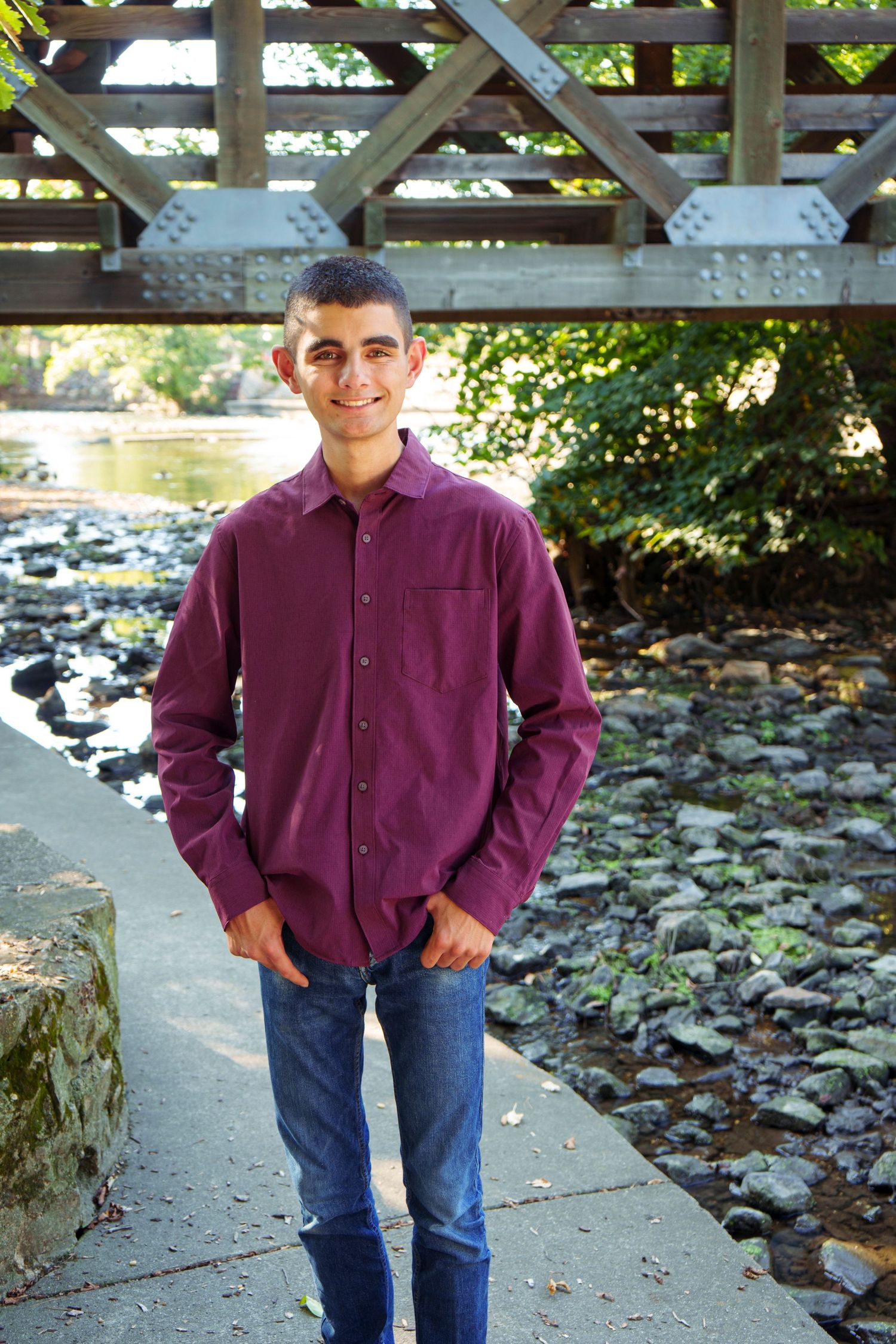 Smiling young man in burgundy shirt standing under wooden bridge near rocky stream.