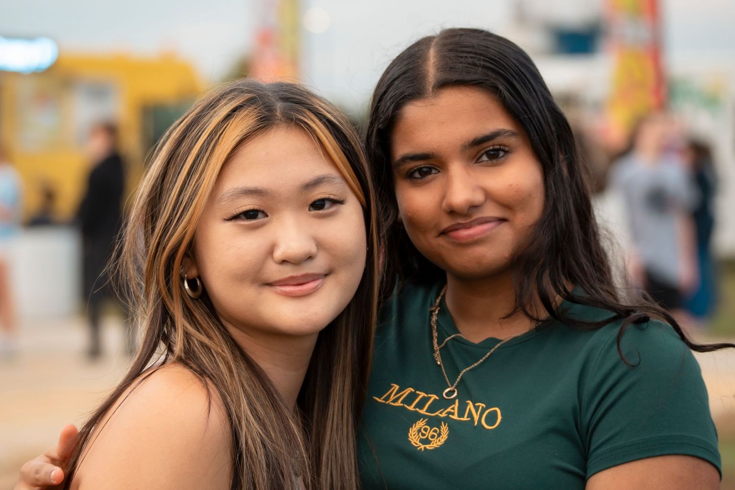 Two teenage girls pose together smiling at an outdoor fair or carnival event.