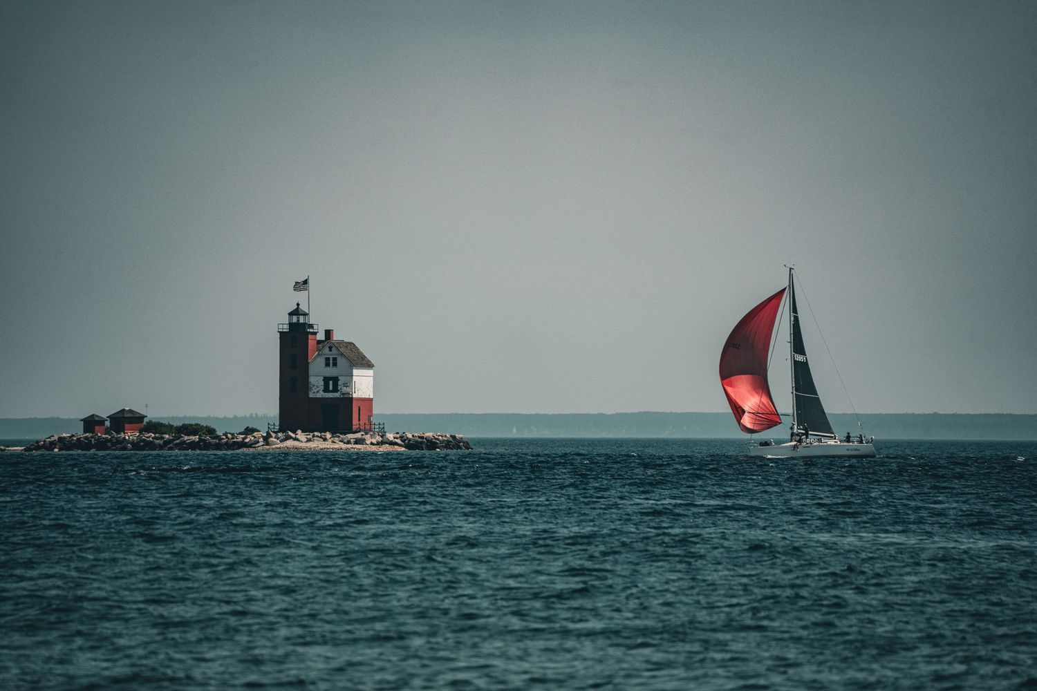 Red sailboat with spinnaker sail passing a historic red lighthouse on a rocky island in calm open water.