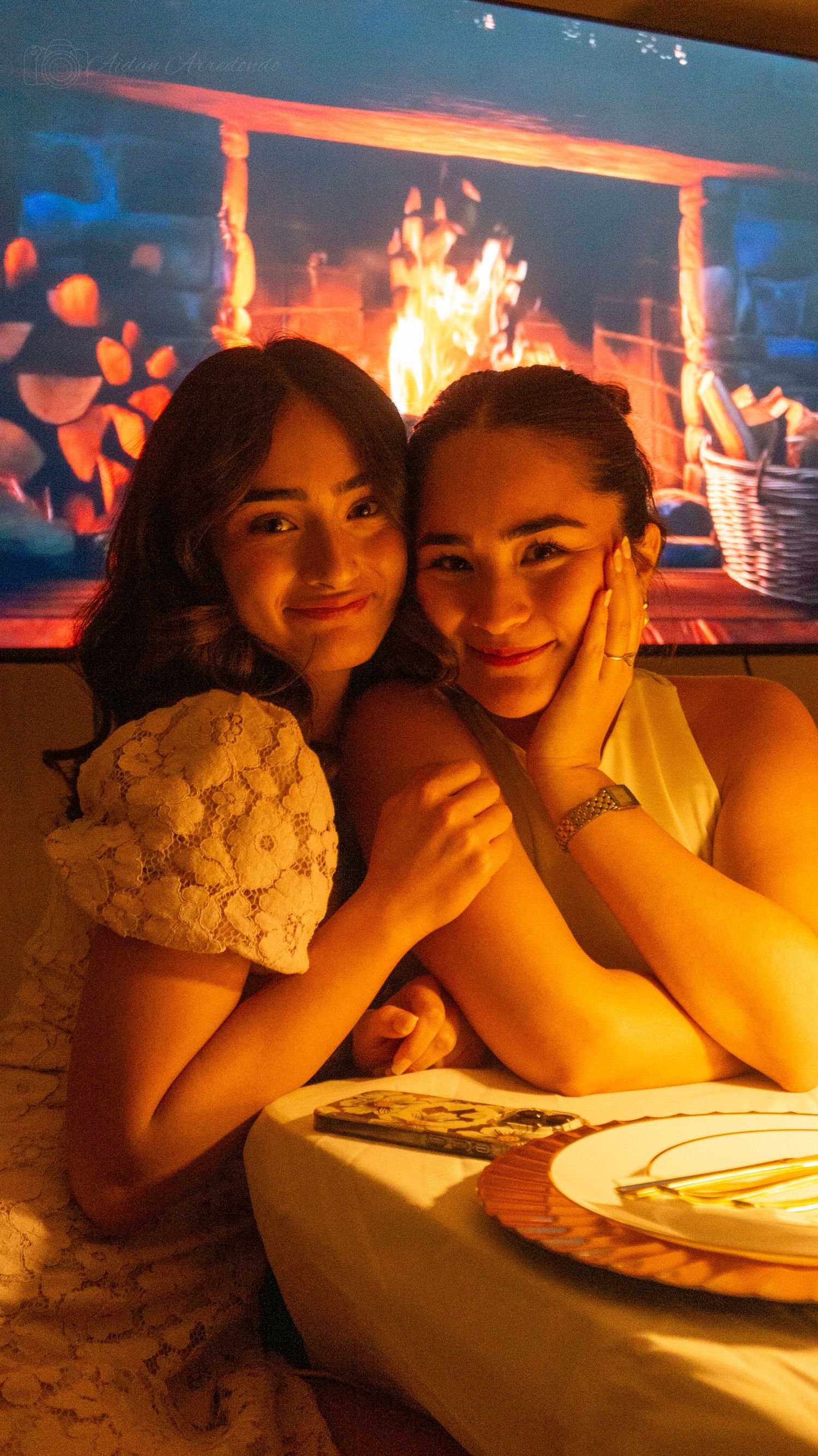 Two young women smiling and embracing at a romantic dinner with a glowing fireplace projection backdrop.